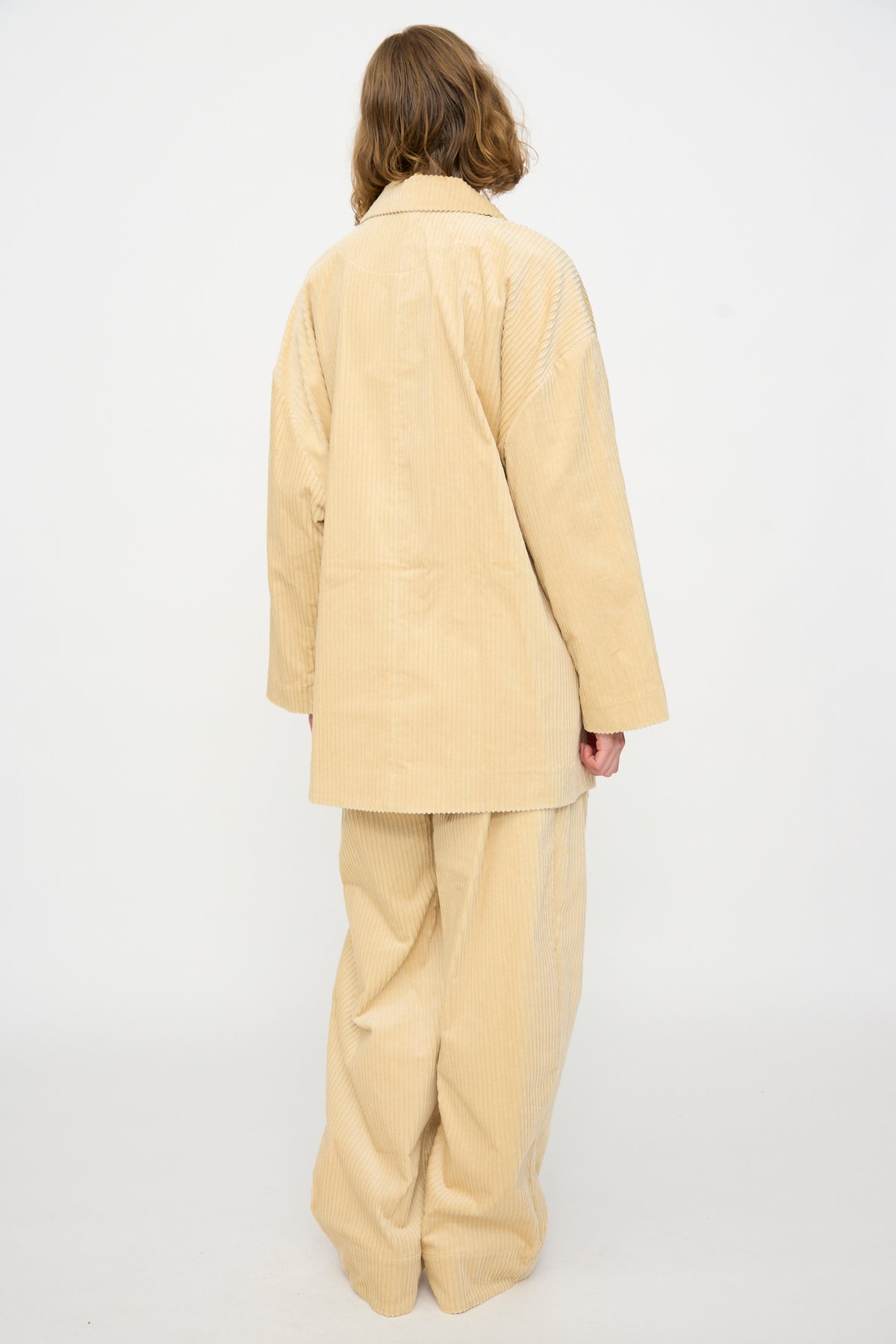 A person with shoulder-length hair stands facing away, wearing the Rachel Comey Chore Jacket in Dune—an oversized beige cotton corduroy piece—paired with wide-legged pants, set against a plain white background.