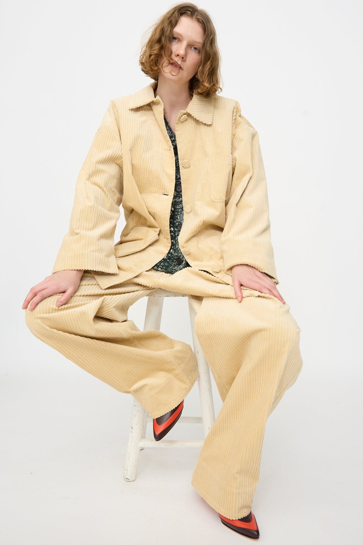A person with wavy hair sits on a white stool, wearing the Rachel Comey Chore Jacket in Dune—an oversized beige cotton corduroy suit—paired with pointed shoes featuring red details, against a plain white background.