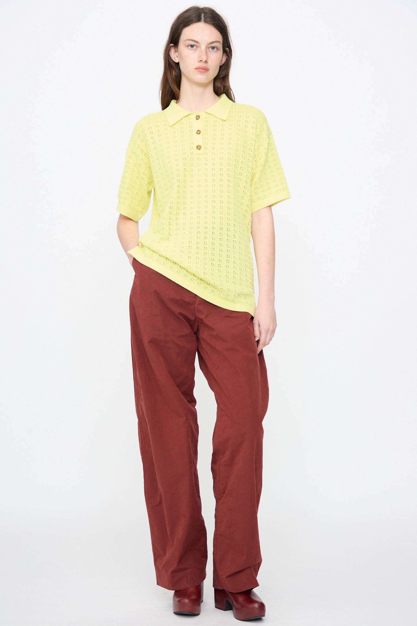 A person stands against a plain white background wearing the Clarence Polo in Limoncello by Rachel Comey, paired with loose maroon pants and maroon shoes.