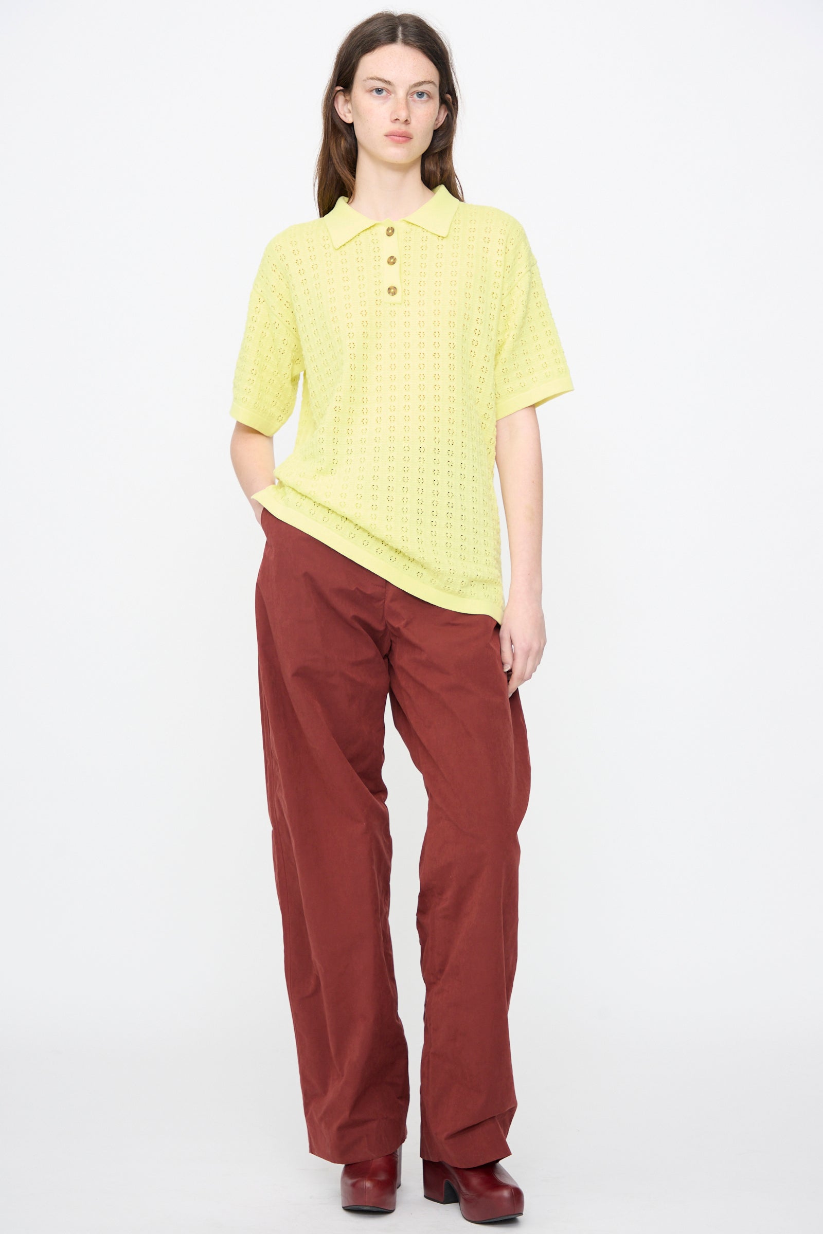 A person stands against a plain white background wearing the Clarence Polo in Limoncello by Rachel Comey, paired with loose maroon pants and maroon shoes.
