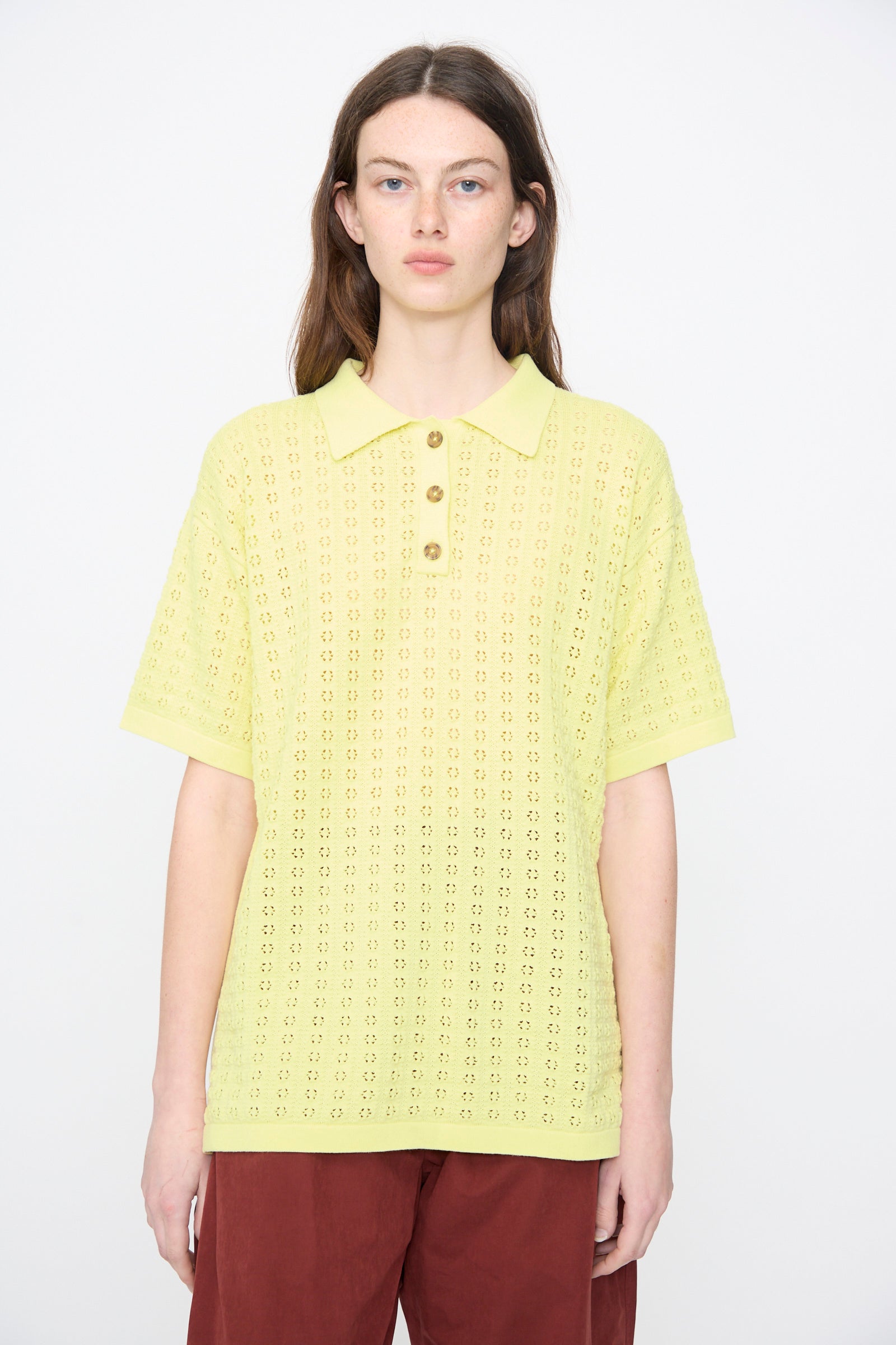 A person stands against a plain background wearing the Rachel Comey Clarence Polo in Limoncello with floral pointelle details, paired with red-brown pants.