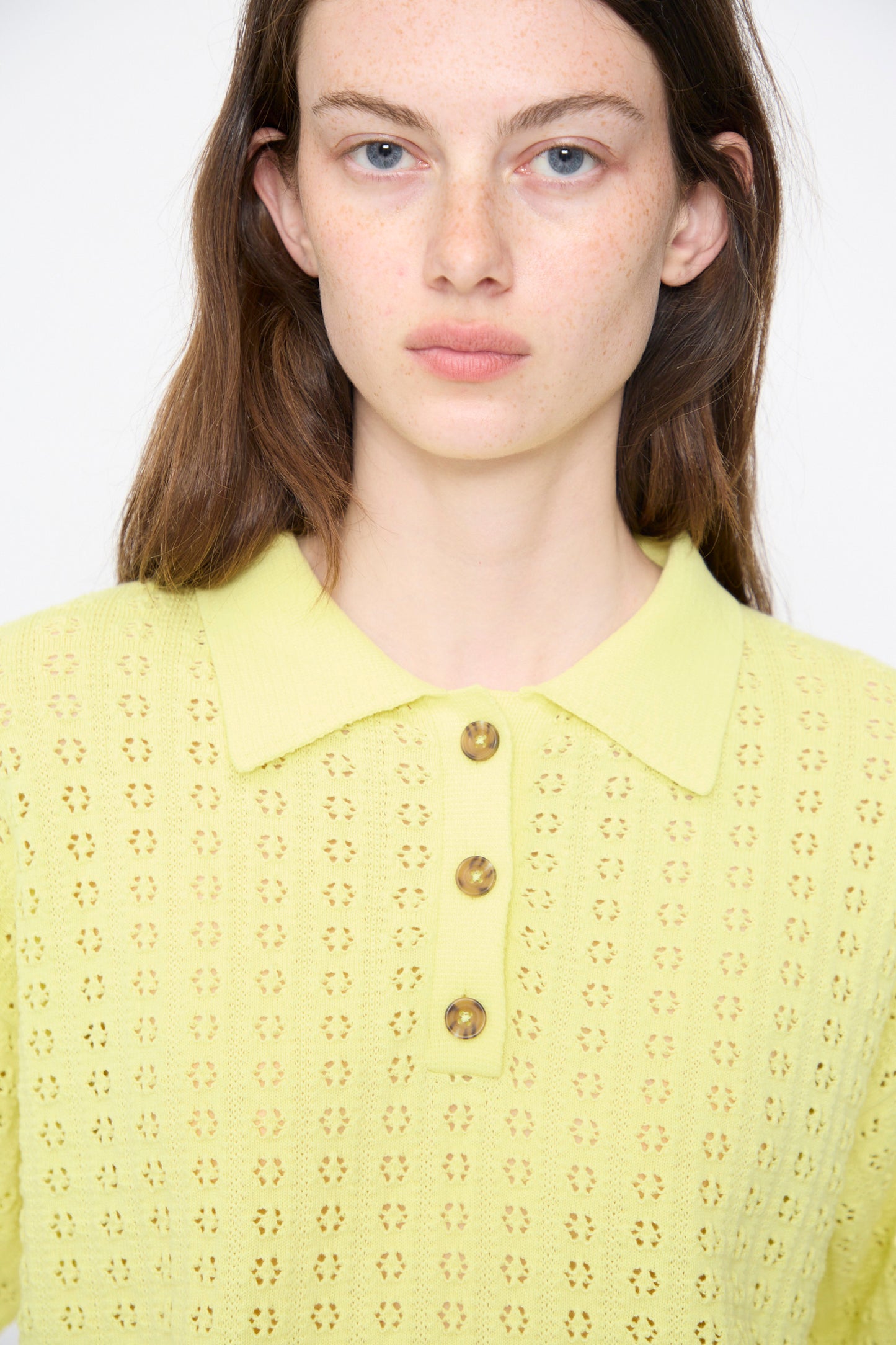 A woman with long brown hair faces the camera against a plain background, wearing the Rachel Comey Clarence Polo in Limoncello—a pale yellow Peruvian cotton shirt with a floral pointelle pattern and buttons.