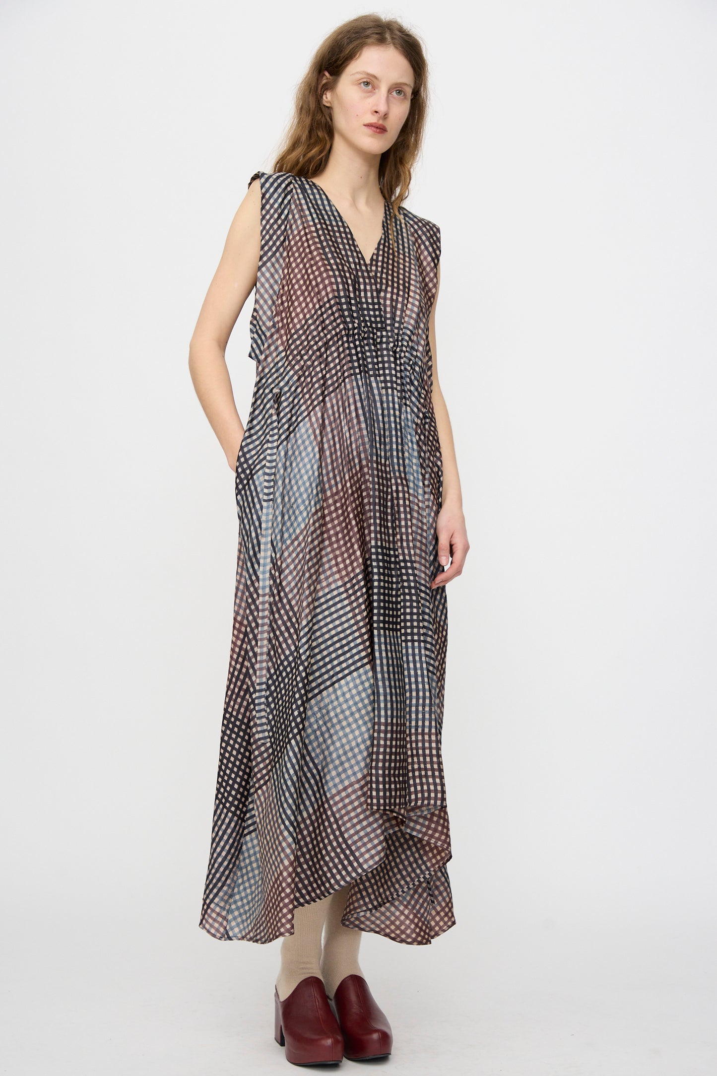 A woman wears the Rachel Comey Clement Dress in Juniper—a silk, sleeveless, V-neck dress with an asymmetric hem—handmade and block printed in India. She pairs it with beige tights and red shoes against a white background.