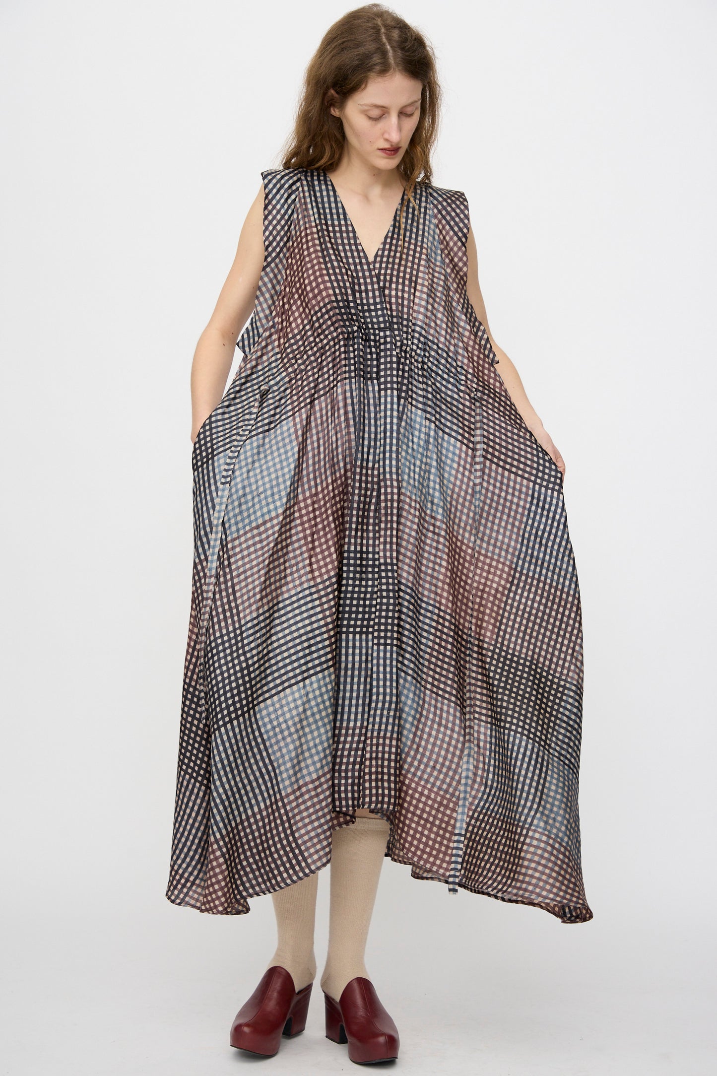 A woman wears the Rachel Comey Clement Dress in Juniper—a loose, block-printed, sleeveless dress handmade in India with a geometric checkered pattern—paired with cream tights and red shoes against a white background.