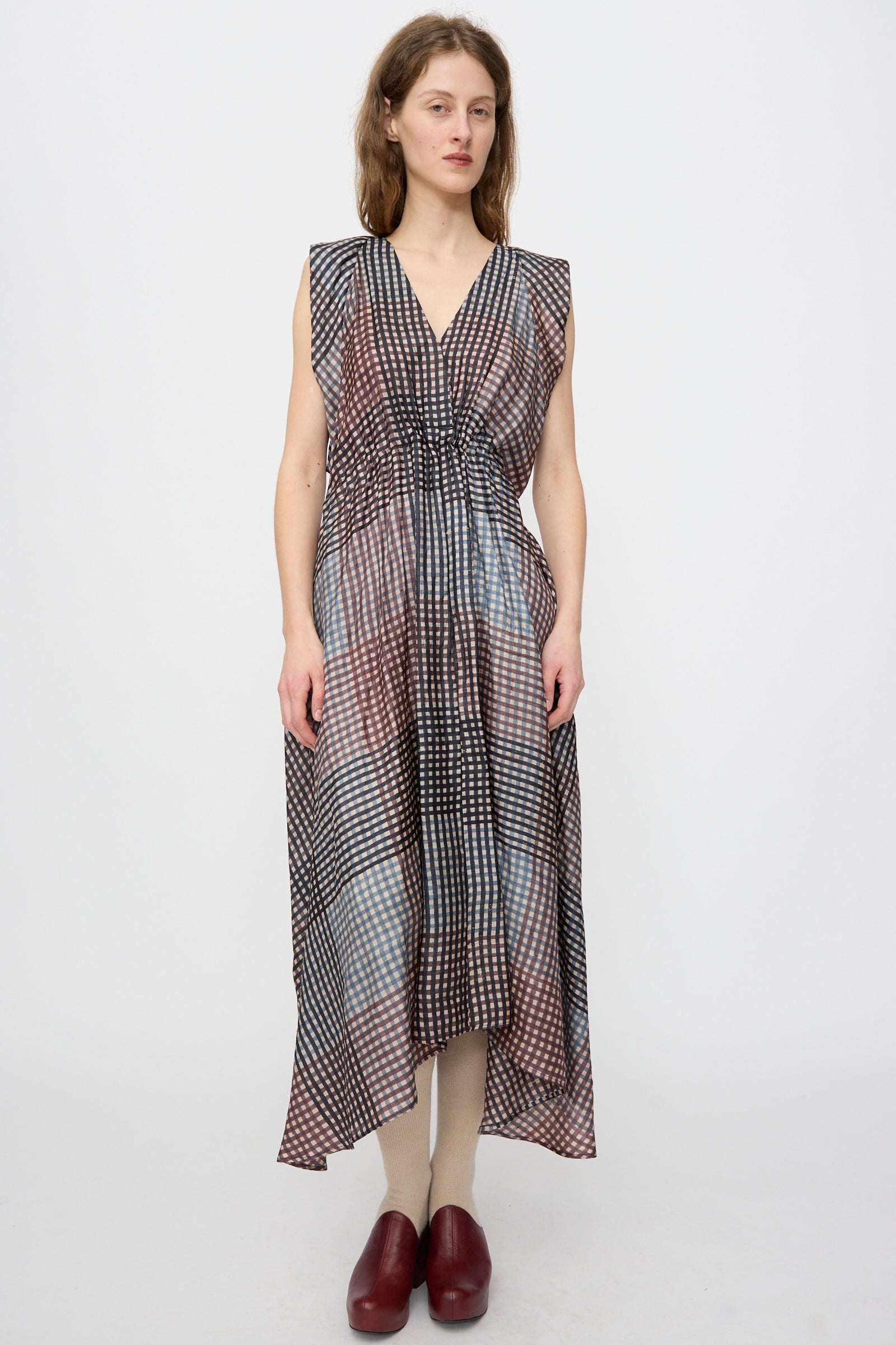 A woman faces forward in the Rachel Comey Clement Dress in Juniper—a silk, sleeveless, V-neck, ankle-length plaid dress—paired with red shoes, handmade in India and photographed against a plain white background.