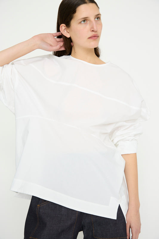 A person with long hair is posing against a plain background, dressed in dark pants and the Rachel Comey Crisp Cotton Lelio Top in white, with one hand behind their head.