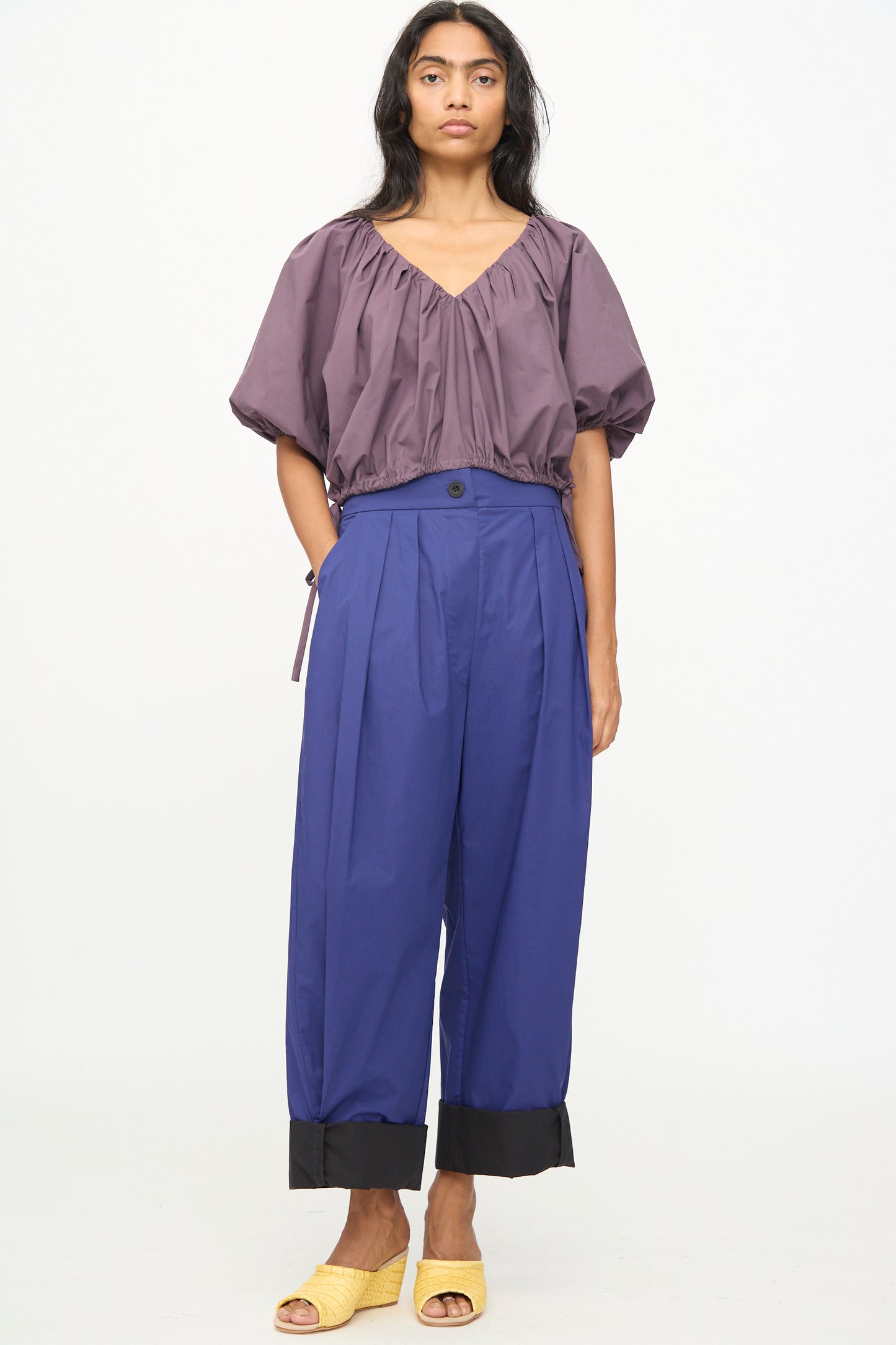 A woman stands against a white background wearing a loose purple top, the Rachel Comey Cuffed Coxsone Pant in blue—a high-waisted, relaxed fit style with wide legs and black cuffs—and yellow open-toe shoes.
