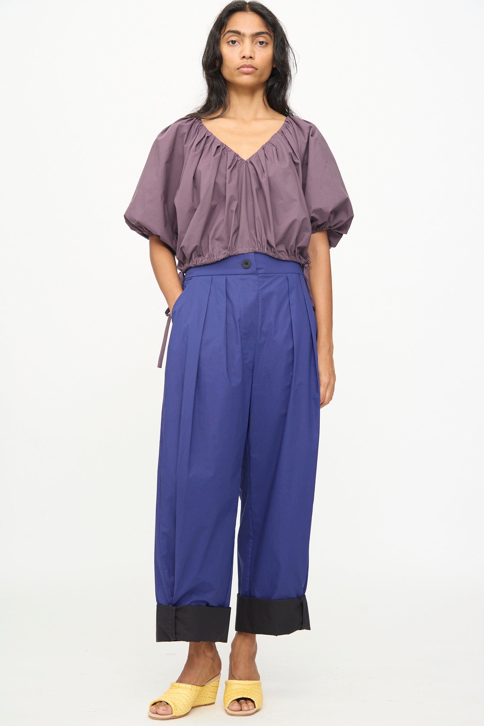 A woman stands against a white background wearing a loose purple top, the Rachel Comey Cuffed Coxsone Pant in blue—a high-waisted, relaxed fit style with wide legs and black cuffs—and yellow open-toe shoes.
