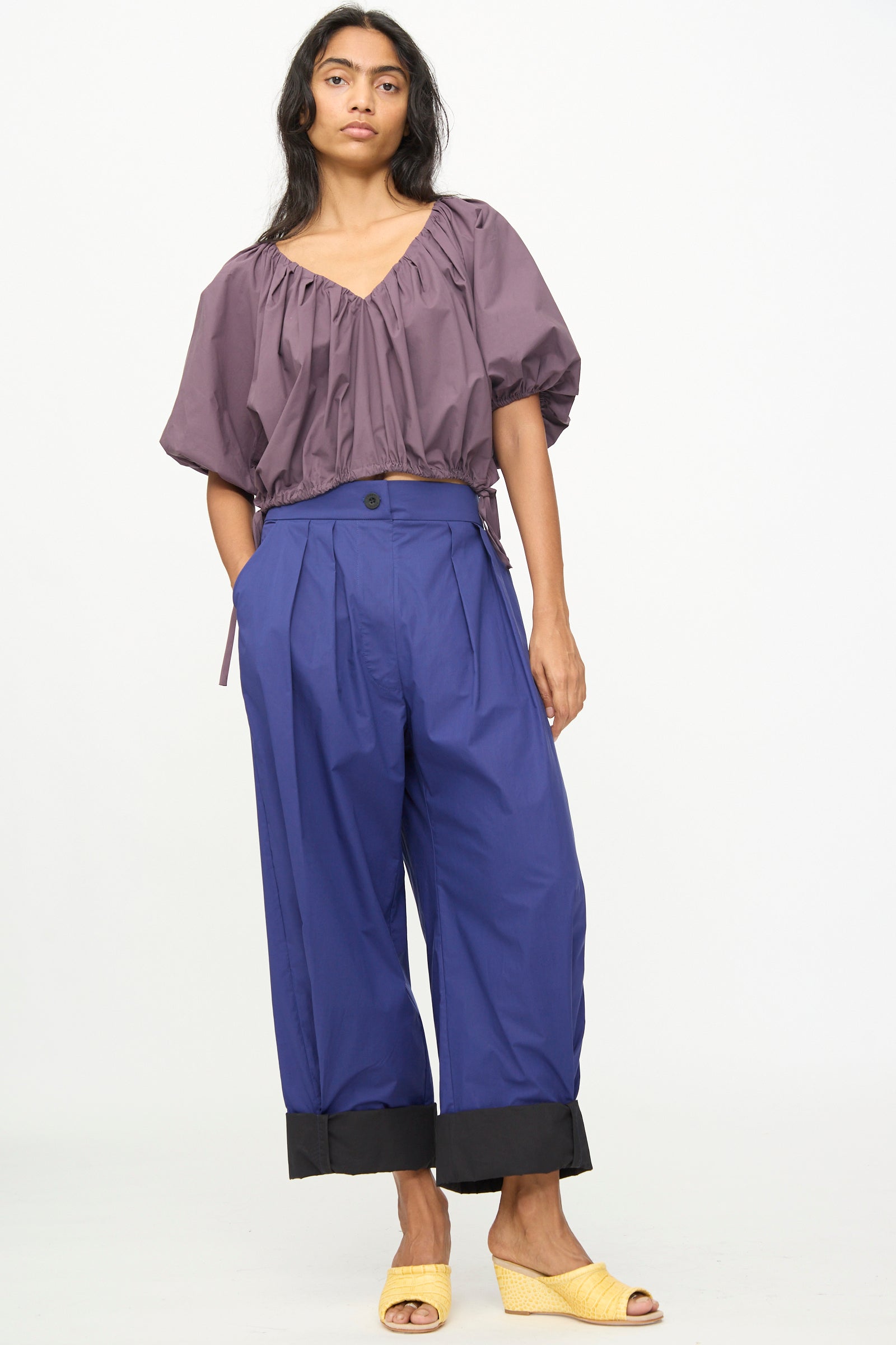 A woman is wearing the Rachel Comey Cuffed Coxsone Pant in Blue—relaxed fit, high-waisted, and pleated with black cuffs—crafted from soft organic cotton. She pairs it with a loose purple blouse and yellow open-toe wedge heels, standing against a plain white background.