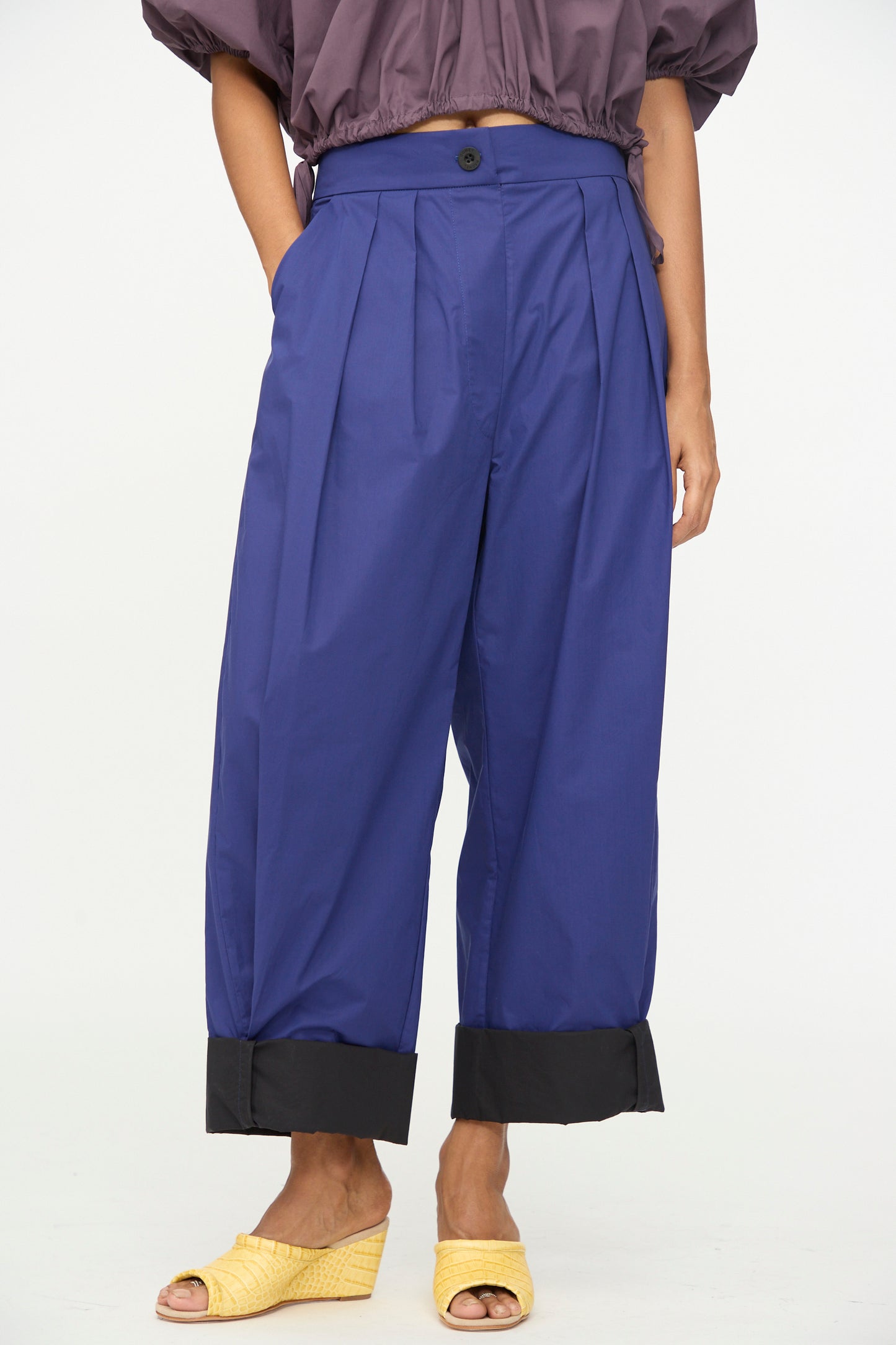 A person wearing the Rachel Comey Cuffed Coxsone Pant in Blue—high-waisted, wide-leg pants with black cuffs and a relaxed fit—paired with a cropped purple top and yellow woven sandals, standing against a plain background.