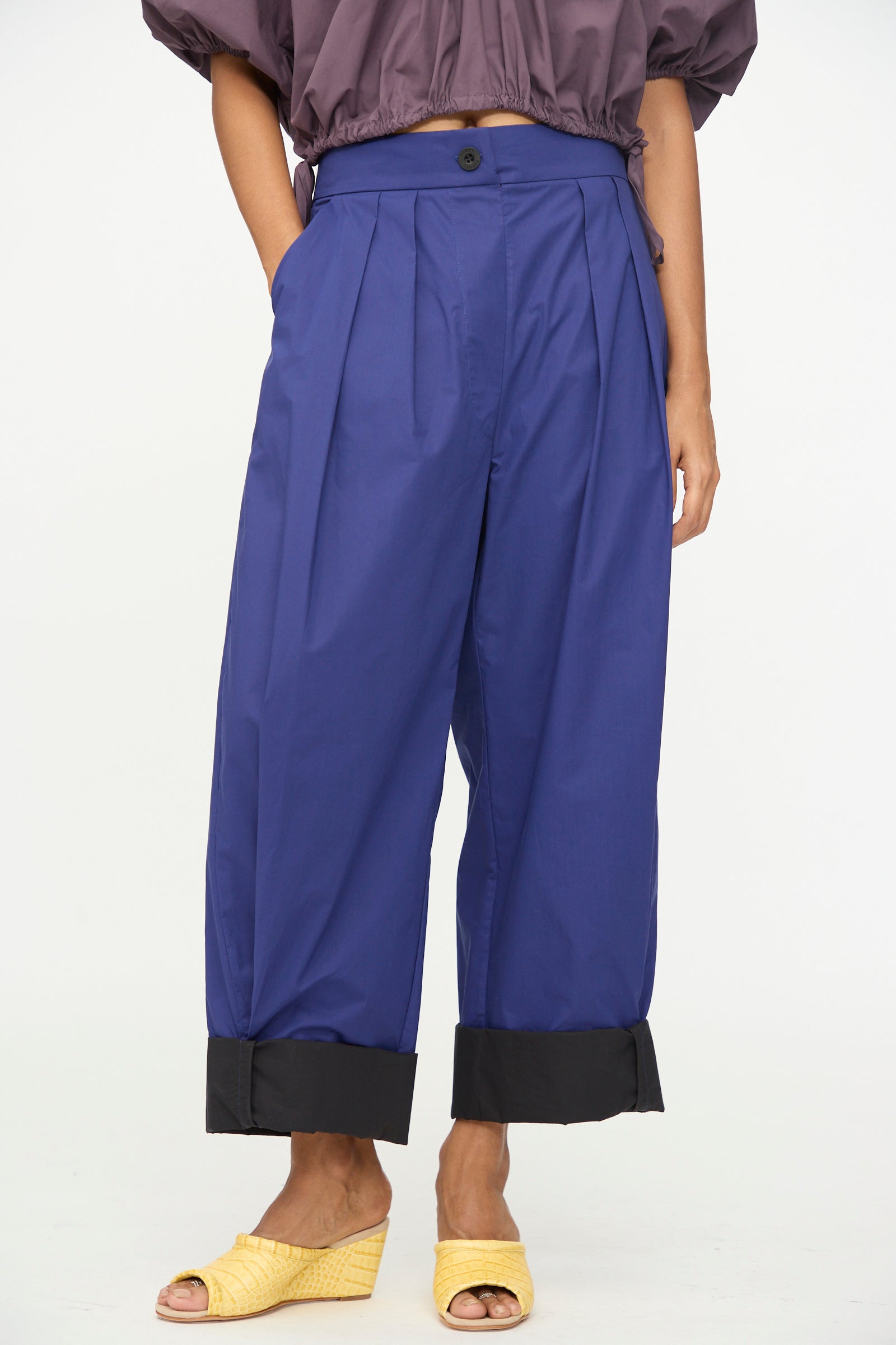 A person wearing the Rachel Comey Cuffed Coxsone Pant in Blue—high-waisted, wide-leg pants with black cuffs and a relaxed fit—paired with a cropped purple top and yellow woven sandals, standing against a plain background.