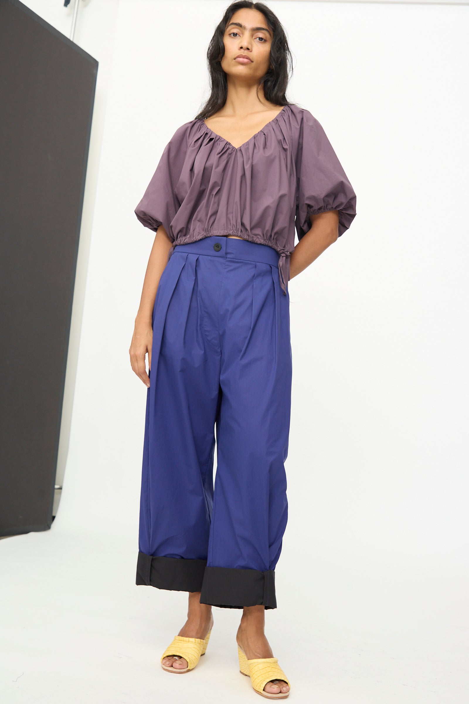 A woman stands against a white background wearing a loose purple top, and the Cuffed Coxsone Pant in Blue by Rachel Comey—an organic cotton, high-waisted pant with a relaxed fit, featuring wide-leg blue legs finished with black cuffs—paired with yellow woven sandals.