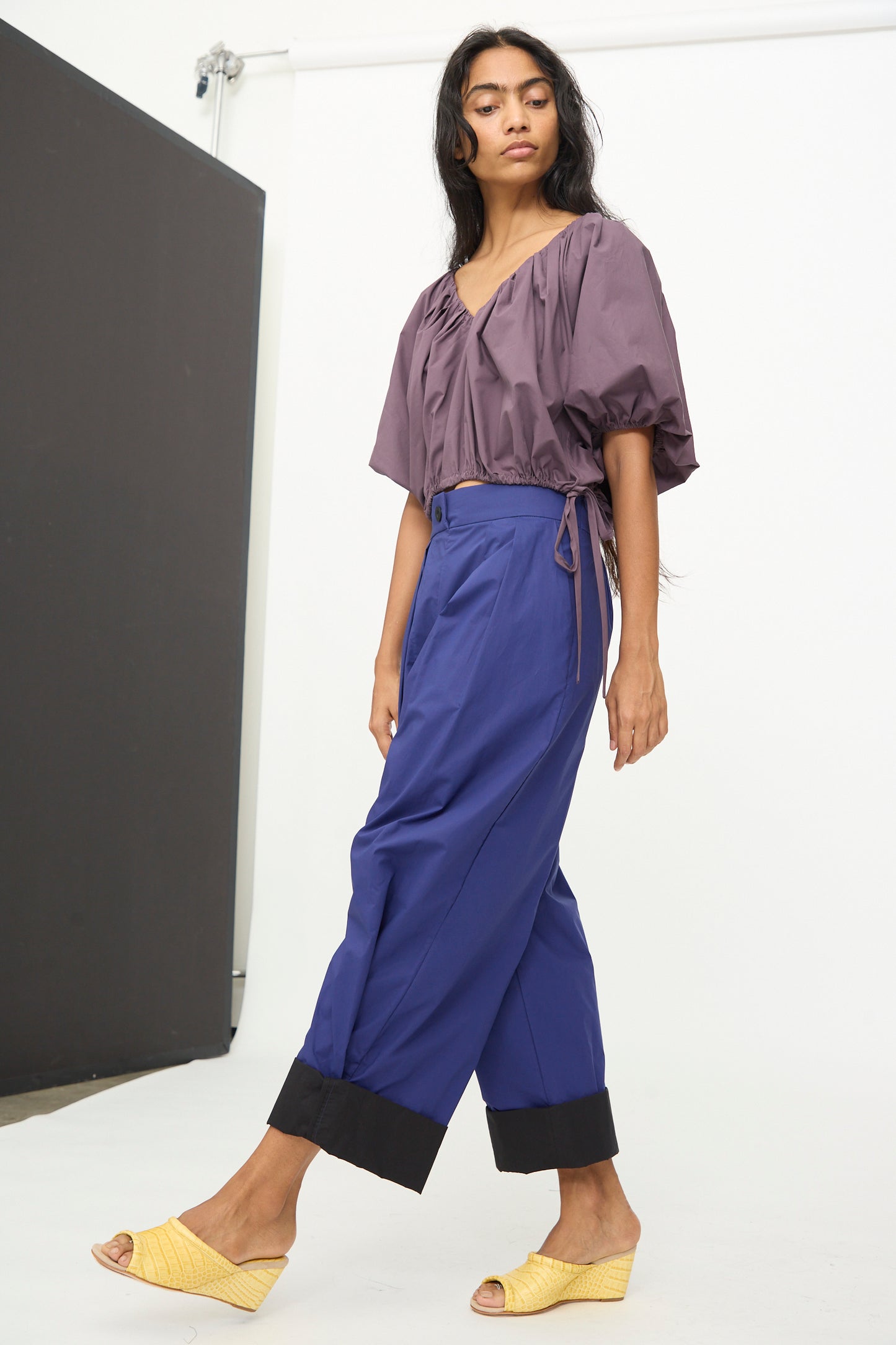 A person stands indoors against a plain background, wearing a loose purple blouse crafted from organic cotton, paired with the Rachel Comey Cuffed Coxsone Pant in Blue and yellow open-toe shoes.