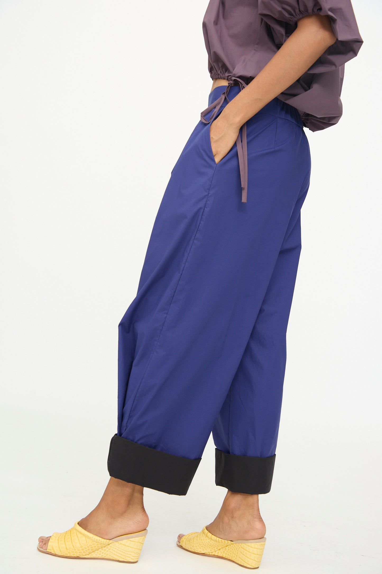A person stands sideways against a plain white background wearing the Rachel Comey Cuffed Coxsone Pant in Blue, a high-waisted, relaxed fit wide-leg pant with black cuffs, paired with a purple blouse and yellow woven wedge sandals.