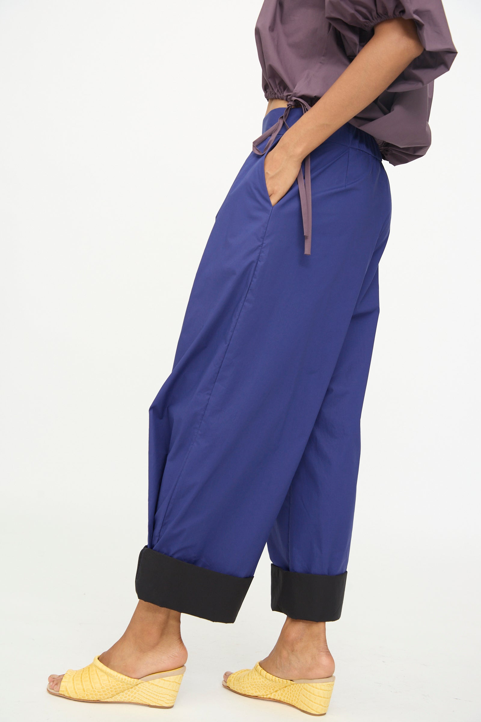 A person stands sideways against a plain white background wearing the Rachel Comey Cuffed Coxsone Pant in Blue, a high-waisted, relaxed fit wide-leg pant with black cuffs, paired with a purple blouse and yellow woven wedge sandals.