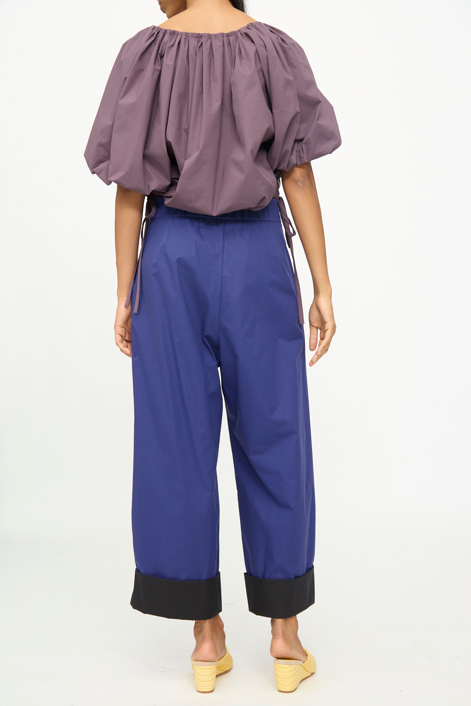 A person stands with their back to the camera, wearing a loose purple top and Rachel Comey’s Cuffed Coxsone Pant in Blue—a relaxed fit, high-waisted wide-leg style featuring black cuffs—paired with yellow heeled sandals, against a plain white background.