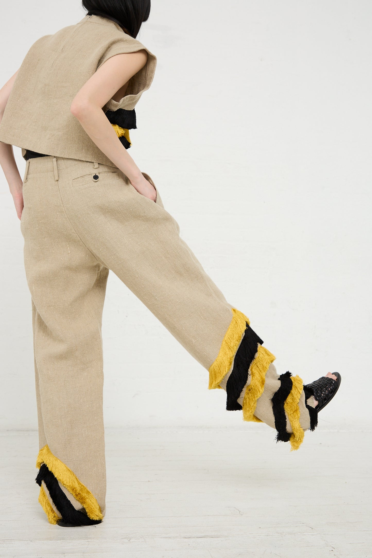 Woman in beige Rachel Comey Ditto Pant in Natural outfit with black and yellow fringed details on textured linen pants, captured mid-step against a white background.