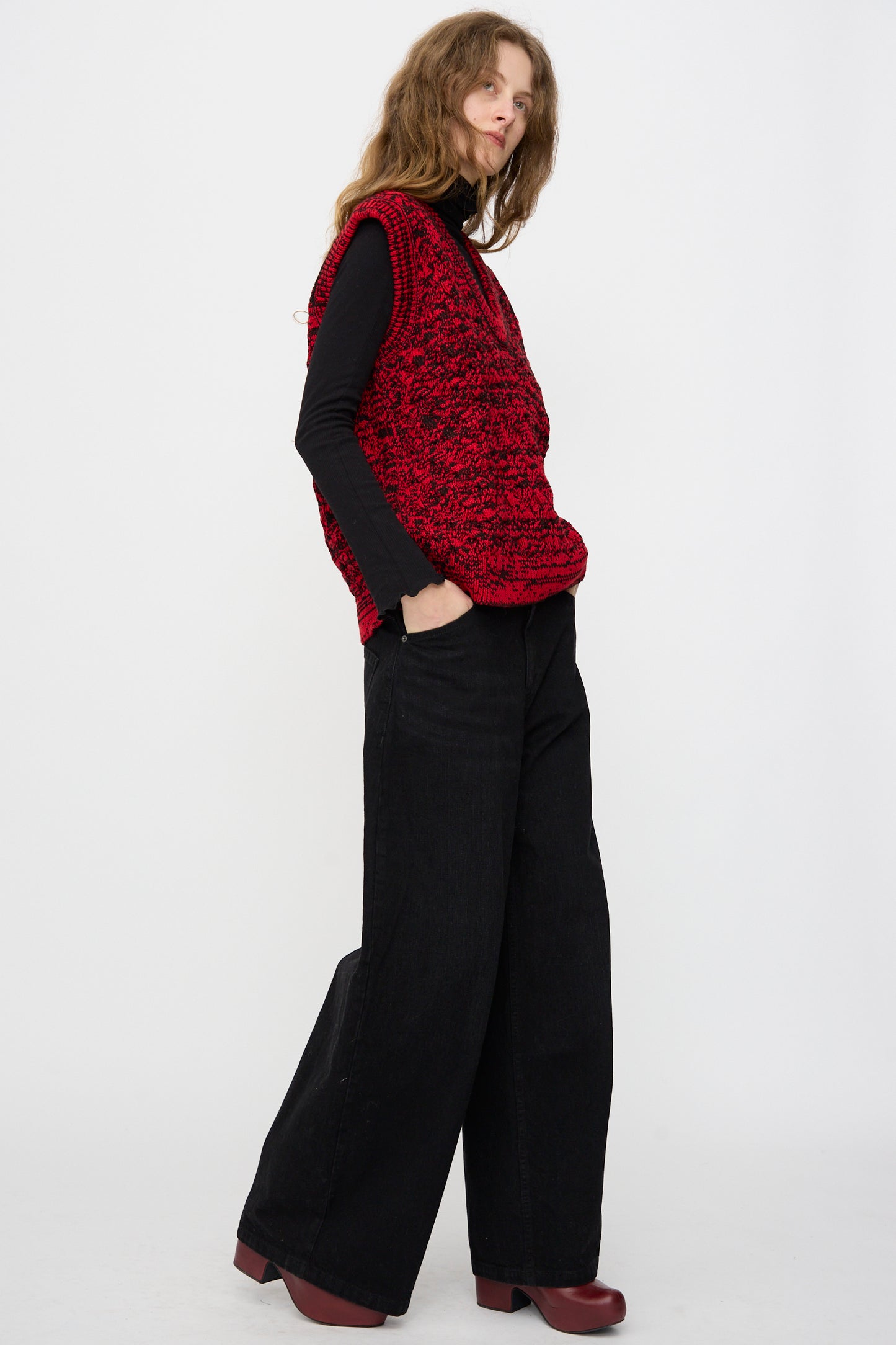 Person wears a textured red vest over a black long-sleeve top, Rachel Comey Dottir Pant in Black, and dark red platform shoes, standing against a plain white background.