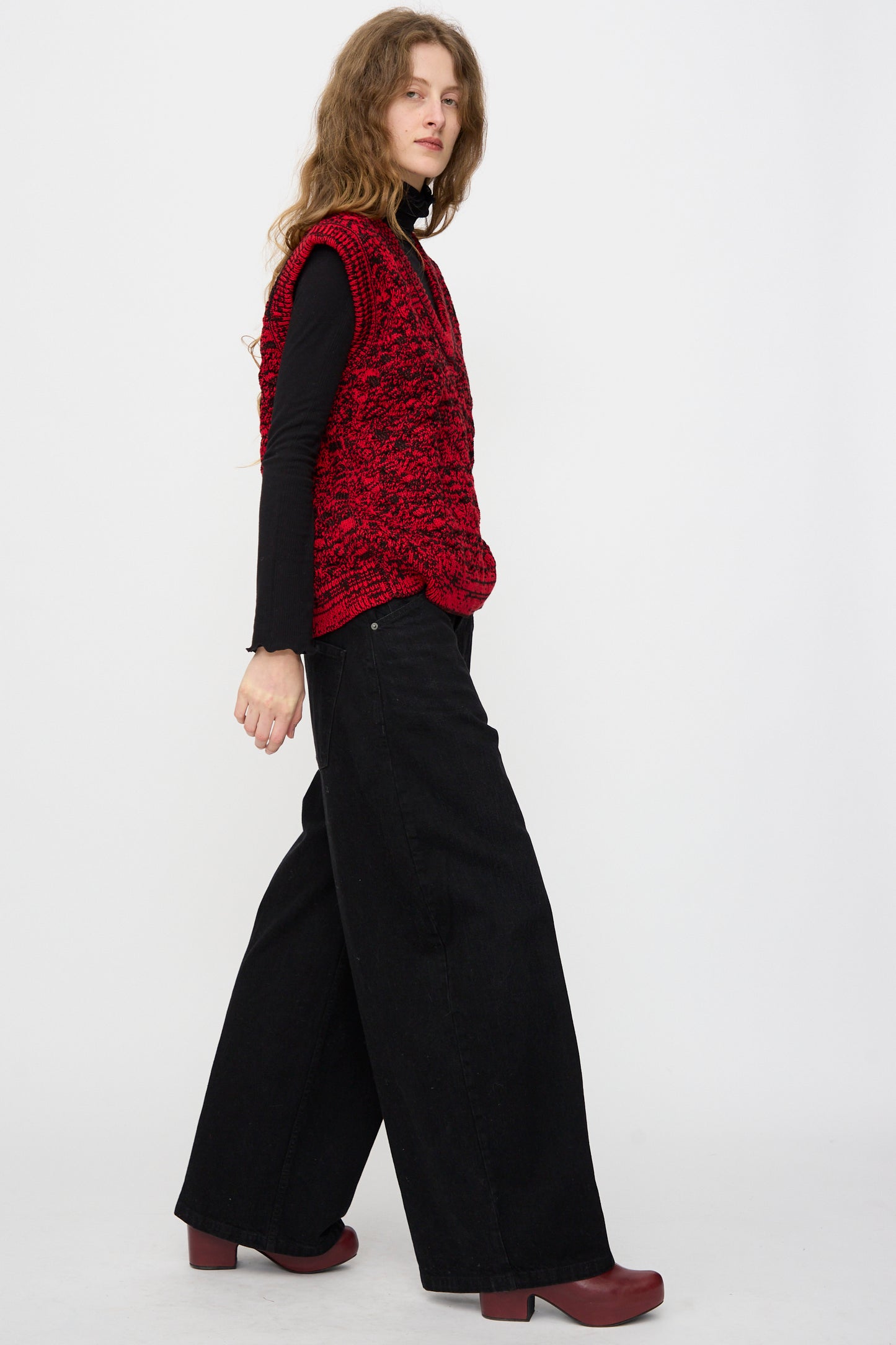 A person stands sideways against a white background, wearing the Rachel Comey Dottir Pant in Black, a red textured vest, black long-sleeve top, and burgundy shoes.