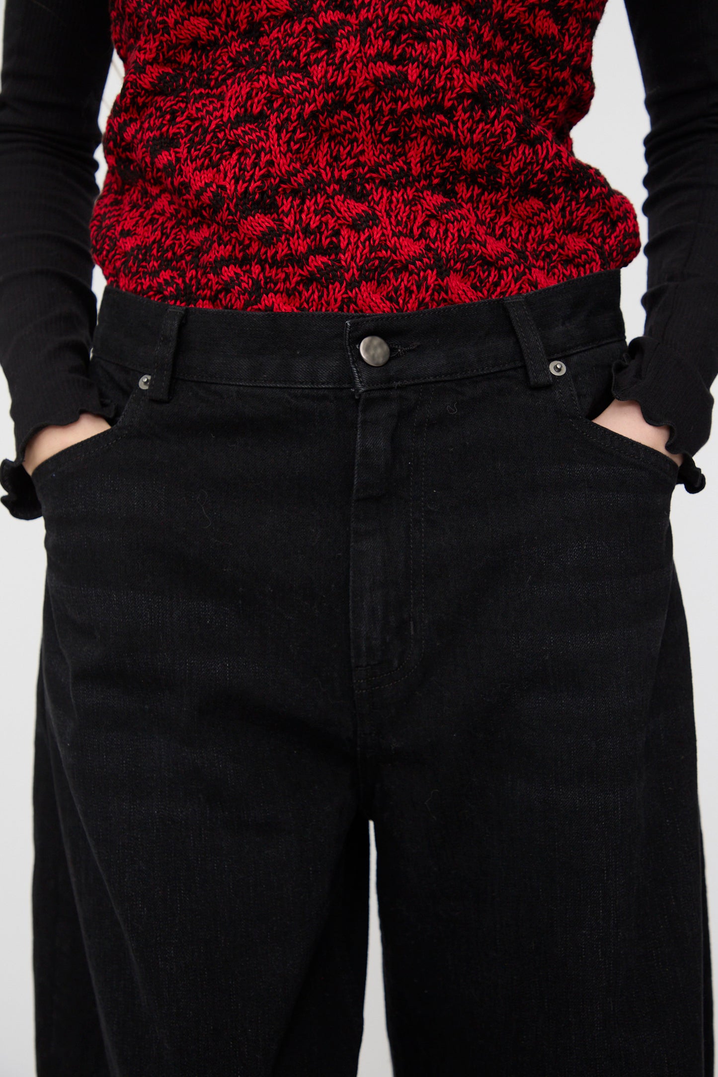 A person wears the Rachel Comey Dottir Pant in Black, pairing the wide-leg denim pants with a red and black patterned top, hands in pockets, photographed from the waist down.