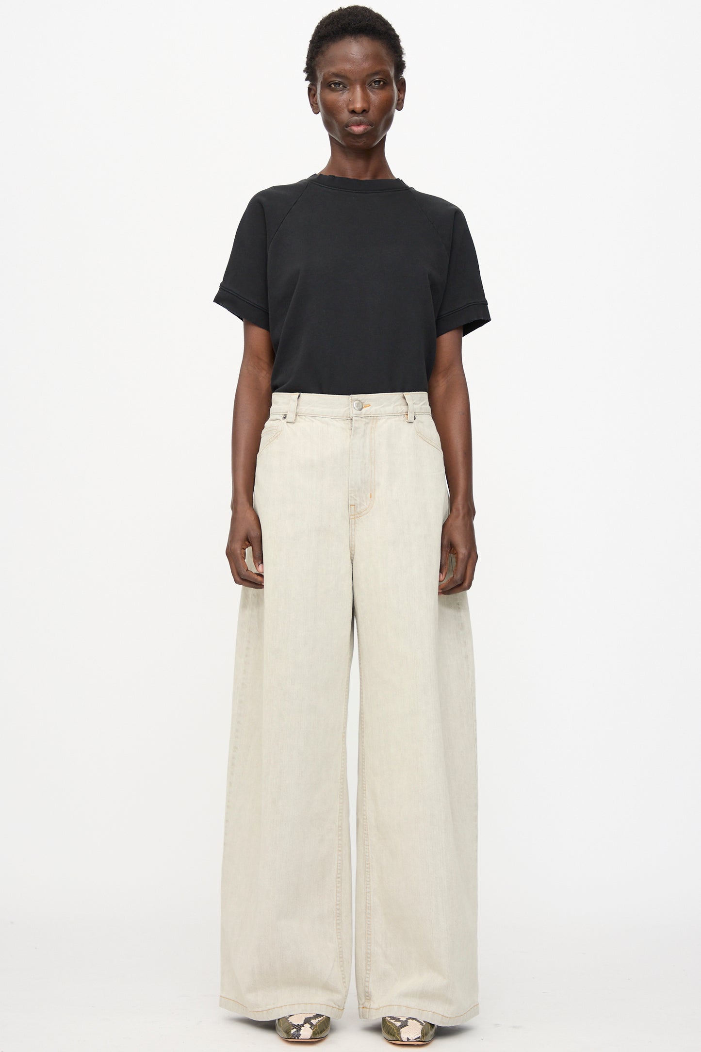 A person stands against a white background wearing a black short-sleeve t-shirt, Rachel Comey Dottir Pant in Khaki Wash, and patterned shoes.