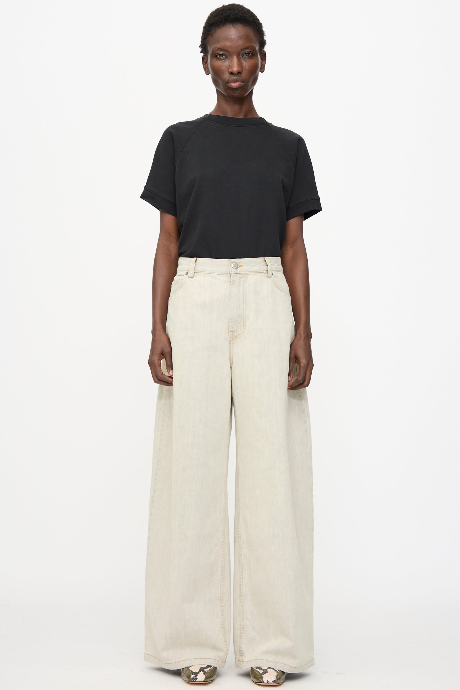 A person stands against a white background wearing a black short-sleeve t-shirt, Rachel Comey Dottir Pant in Khaki Wash, and patterned shoes.