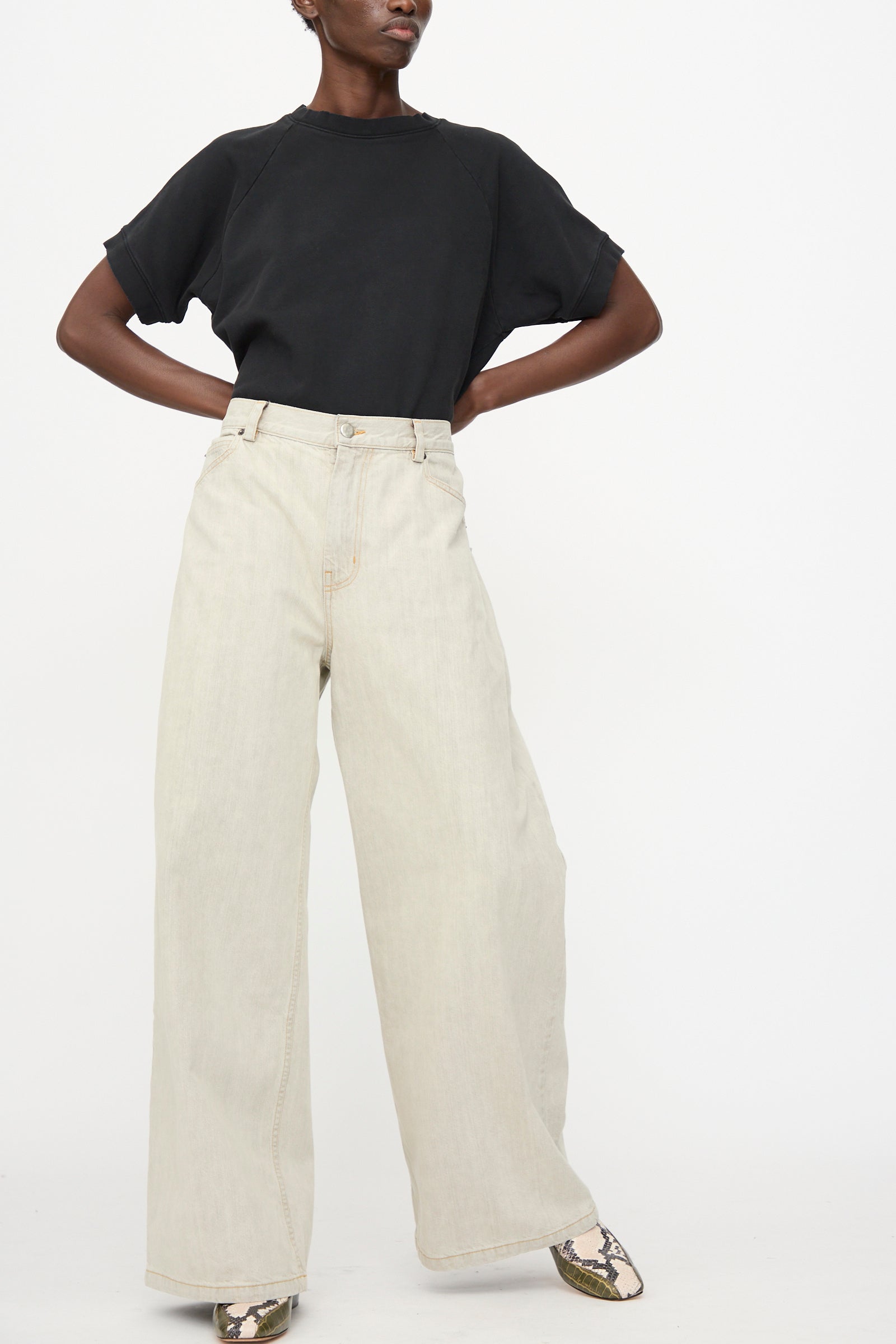 A person wearing a black short-sleeve top, Rachel Comey Dottir Pant in Khaki Wash, and snakeskin-patterned shoes stands against a plain white background.