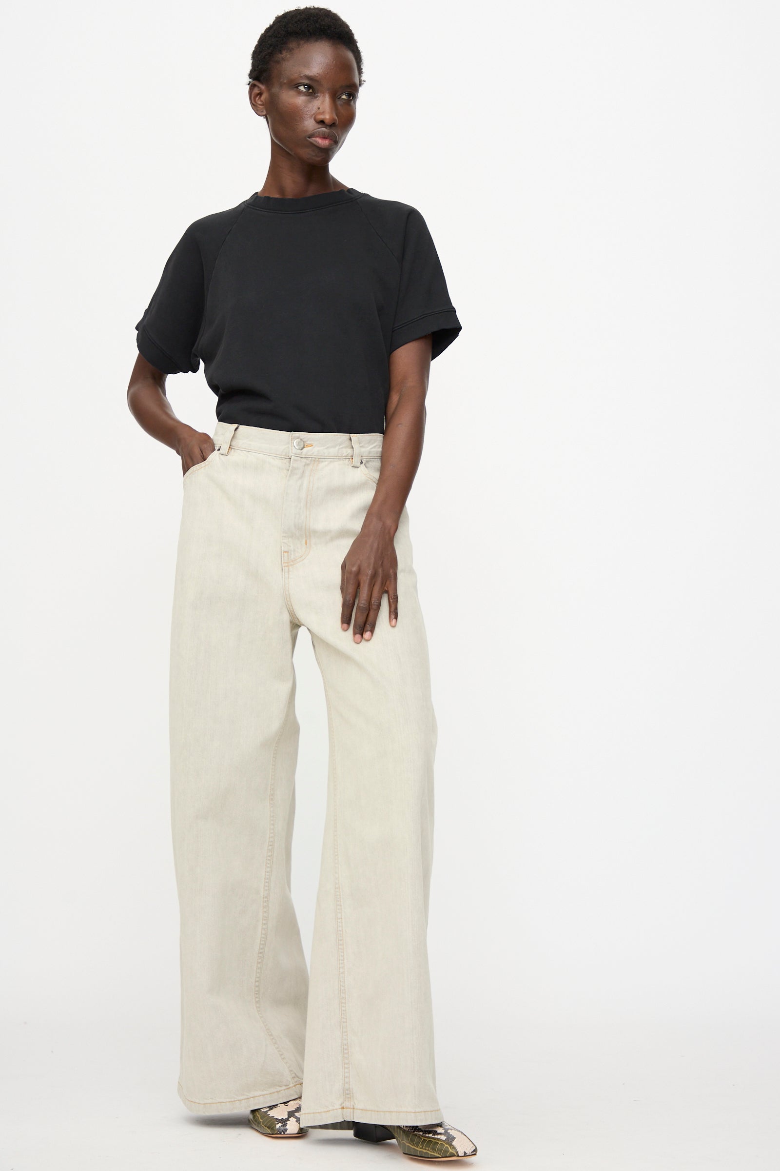 A person stands against a plain background wearing a black short-sleeved t-shirt, Rachel Comey Dottir Pant in Khaki Wash, and patterned shoes.
