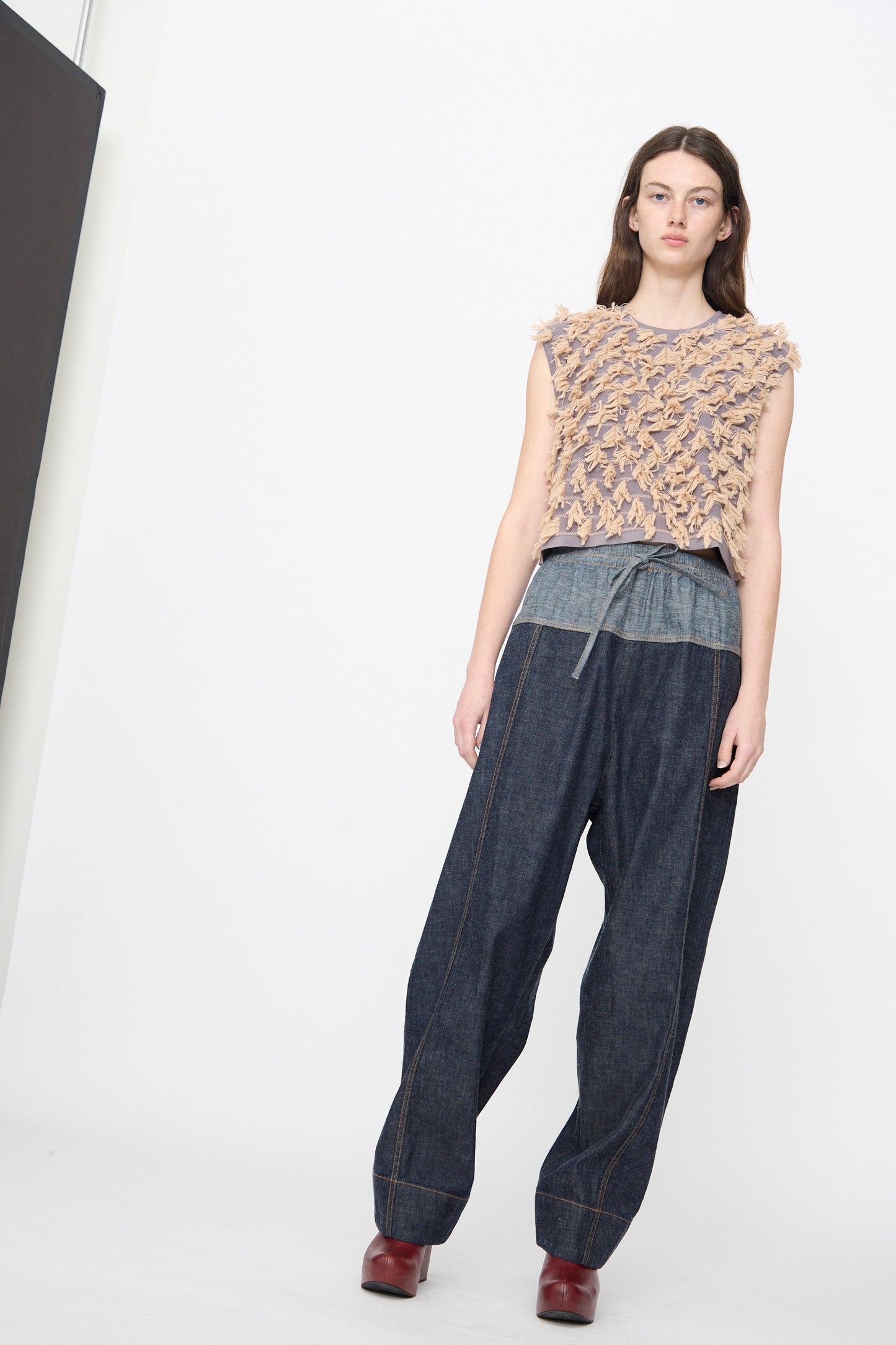 Woman stands against a white background wearing the Ennu Pant in Indigo by Rachel Comey, paired with a textured sleeveless top, two-tone denim shirt, and red shoes.