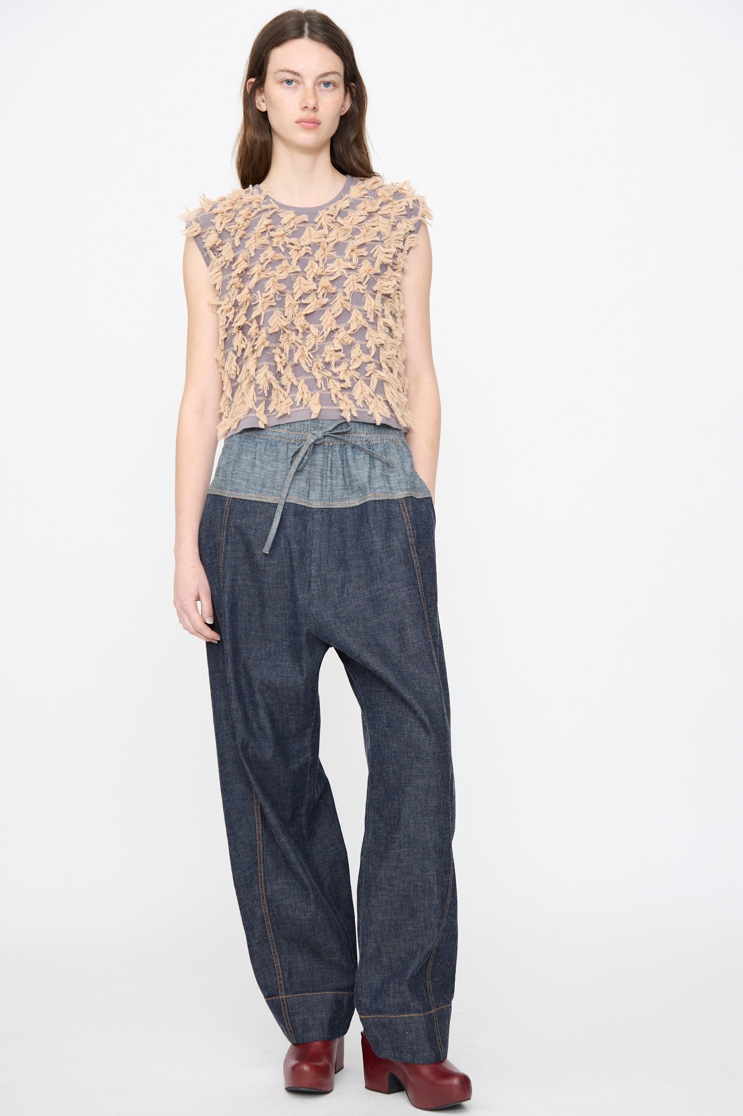 A person stands against a plain background wearing the Rachel Comey Ennu Pant in Indigo, styled with a textured sleeveless beige top, layered shirt, and red heeled shoes.