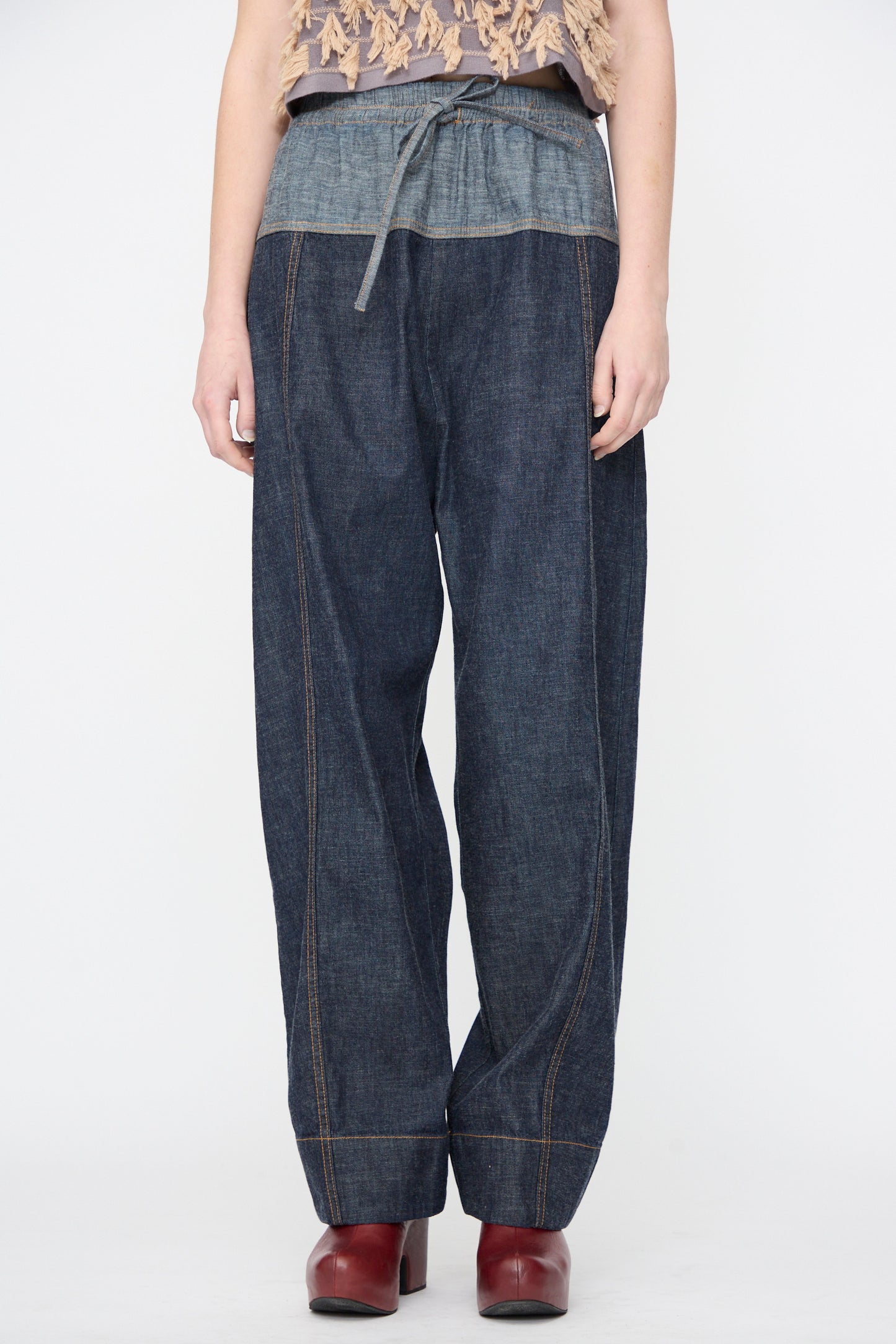 A person wearing the Rachel Comey Ennu Pant in Indigo, featuring a relaxed fit and drawstring waist, paired with red heeled shoes, stands against a plain white background.