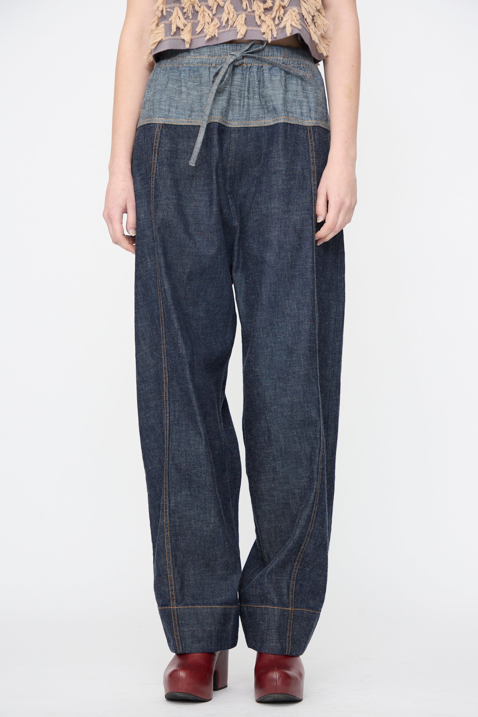A person wearing the Rachel Comey Ennu Pant in Indigo, featuring a relaxed fit and drawstring waist, paired with red heeled shoes, stands against a plain white background.