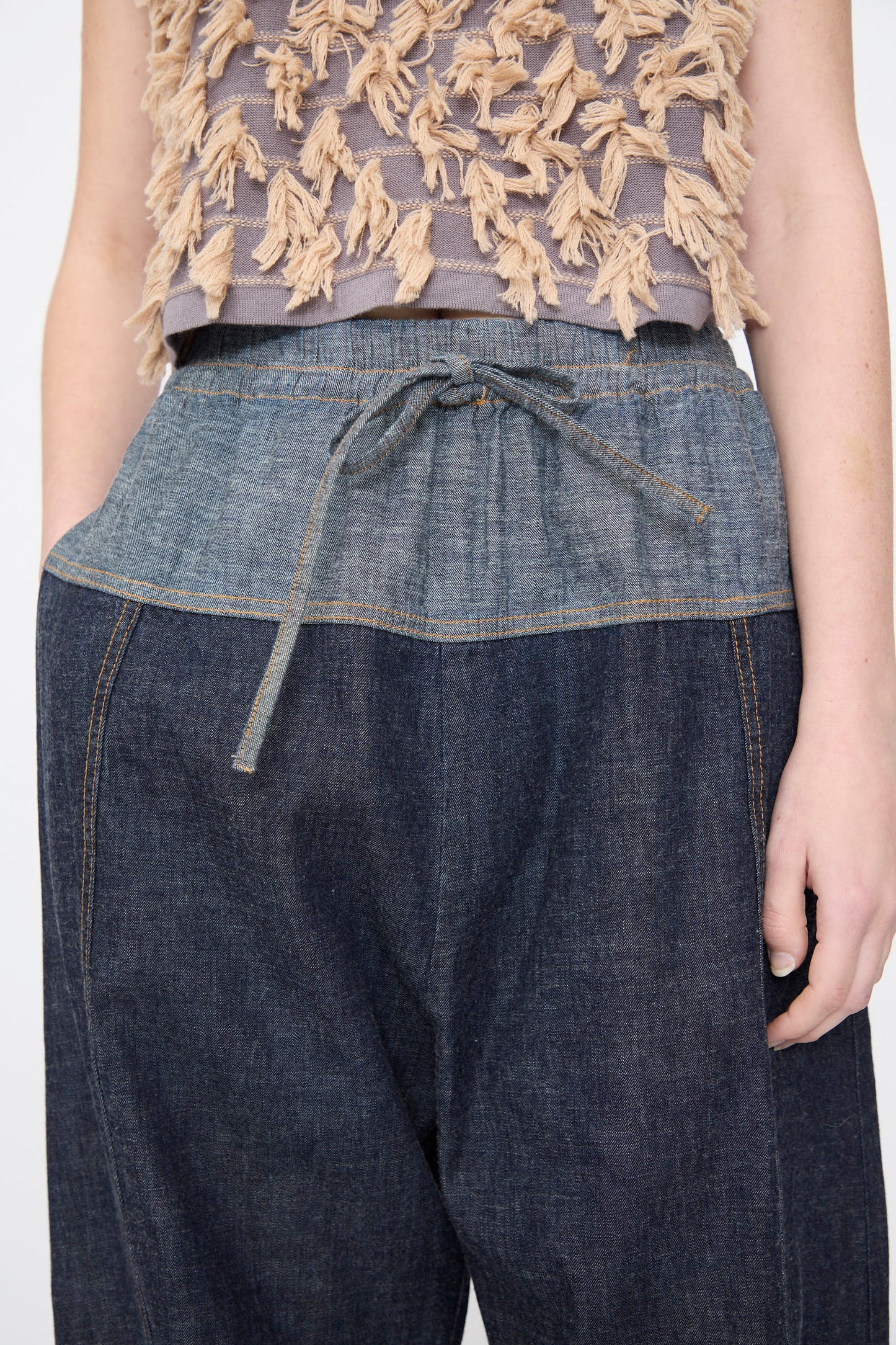 A person wearing Rachel Comey’s Ennu Pant in Indigo, a relaxed-fit dark denim style with a drawstring waist, paired with a sleeveless beige and gray textured top.