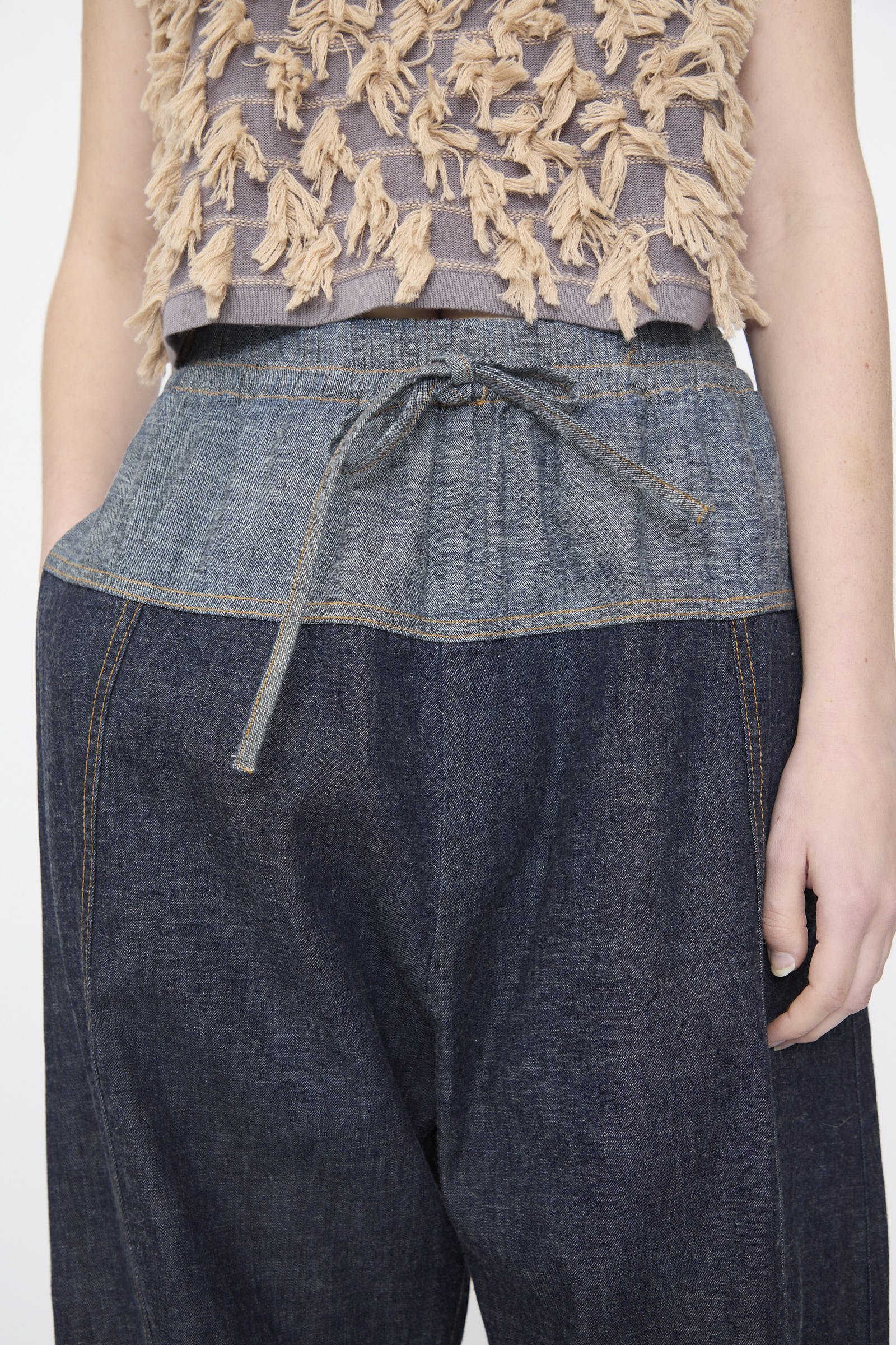 A person wearing Rachel Comey’s Ennu Pant in Indigo, a relaxed-fit dark denim style with a drawstring waist, paired with a sleeveless beige and gray textured top.