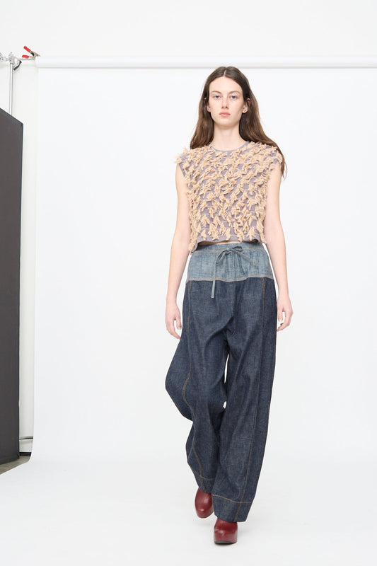 A model walks on a white runway wearing a textured beige sleeveless top, Rachel Comey Ennu Pant in Indigo with a tie waist, and burgundy shoes.