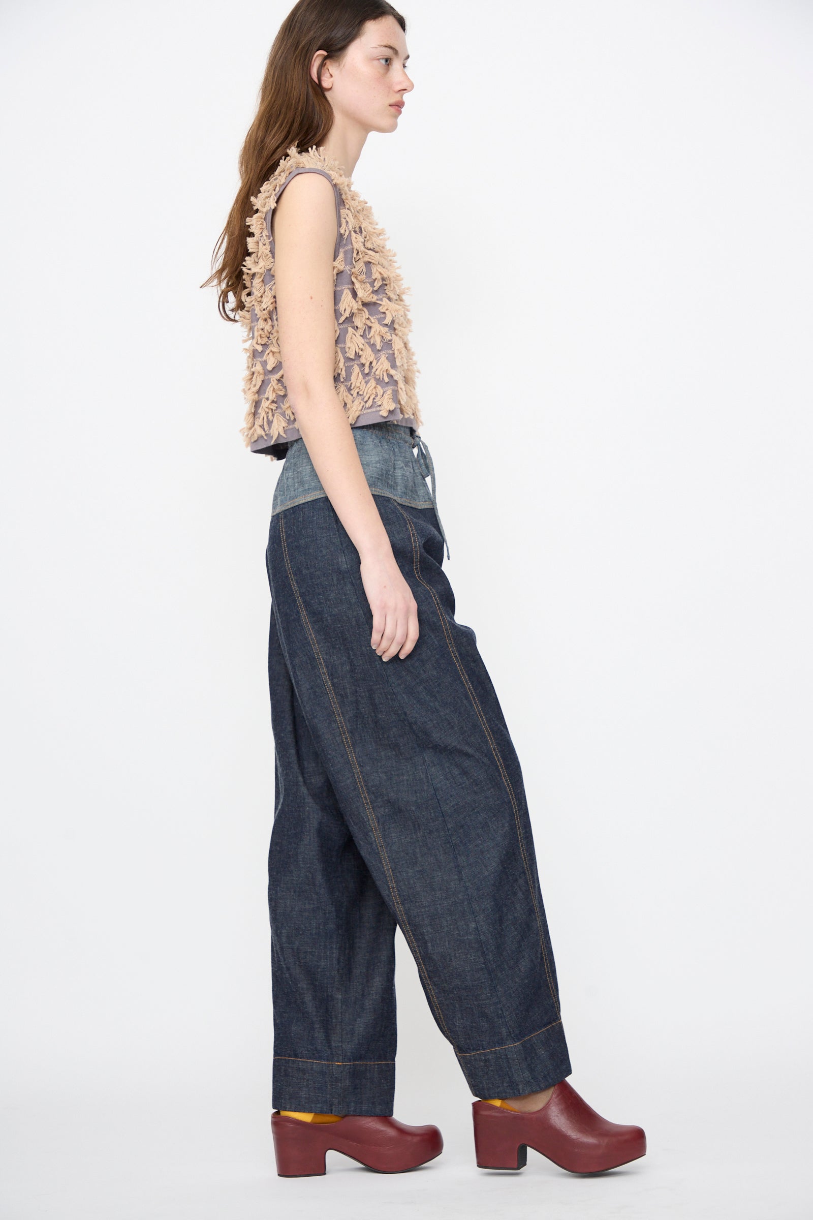 A woman stands in profile against a white background wearing a textured sleeveless top, maroon platform clogs, and the Rachel Comey Ennu Pant in Indigo—relaxed-fit, wide-leg organic cotton denim.