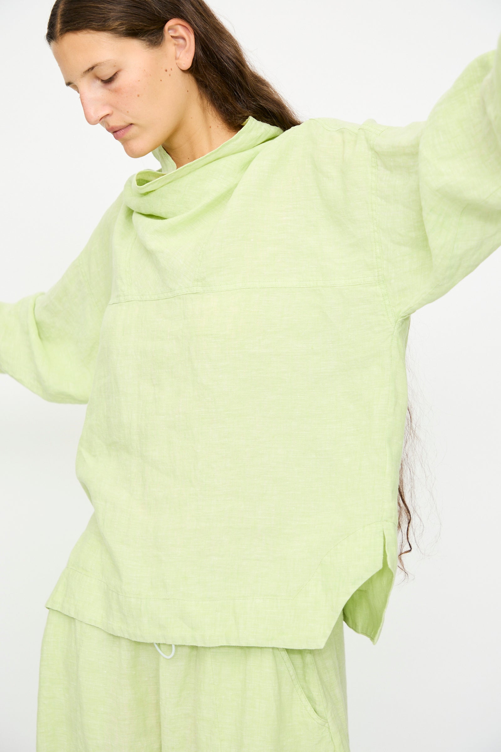 A person wears the Rachel Comey Fiamma Top in White Sage, featuring a lightweight linen, relaxed fit and long sleeves, paired with matching pants, standing with arms slightly raised against a plain white background.