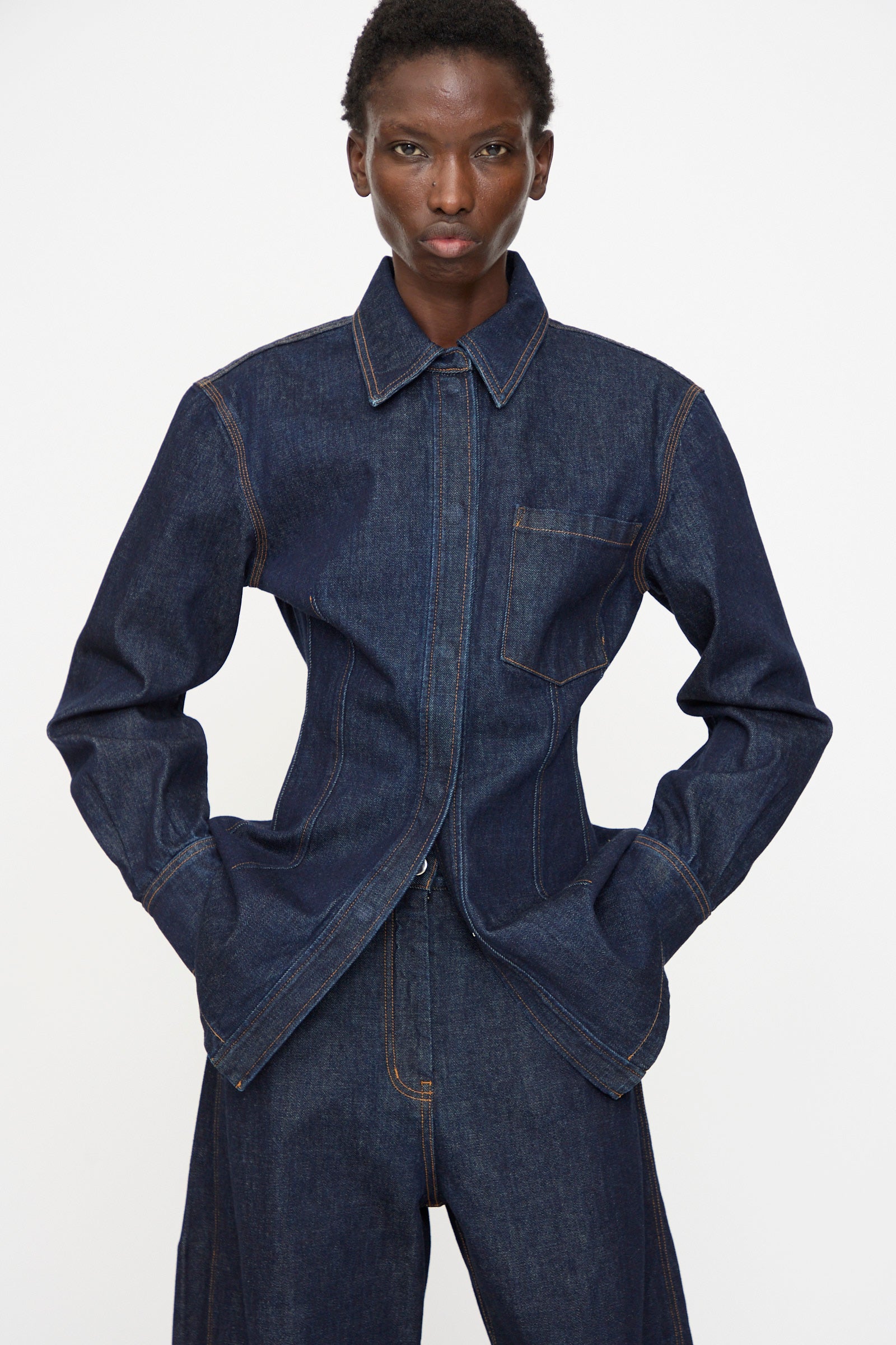 A person wears the Rachel Comey Filt Top in Raw Wash—a dark denim shirt—layered under a matching jacket and pants with tan stitching, standing against a plain white background with hands in pockets.