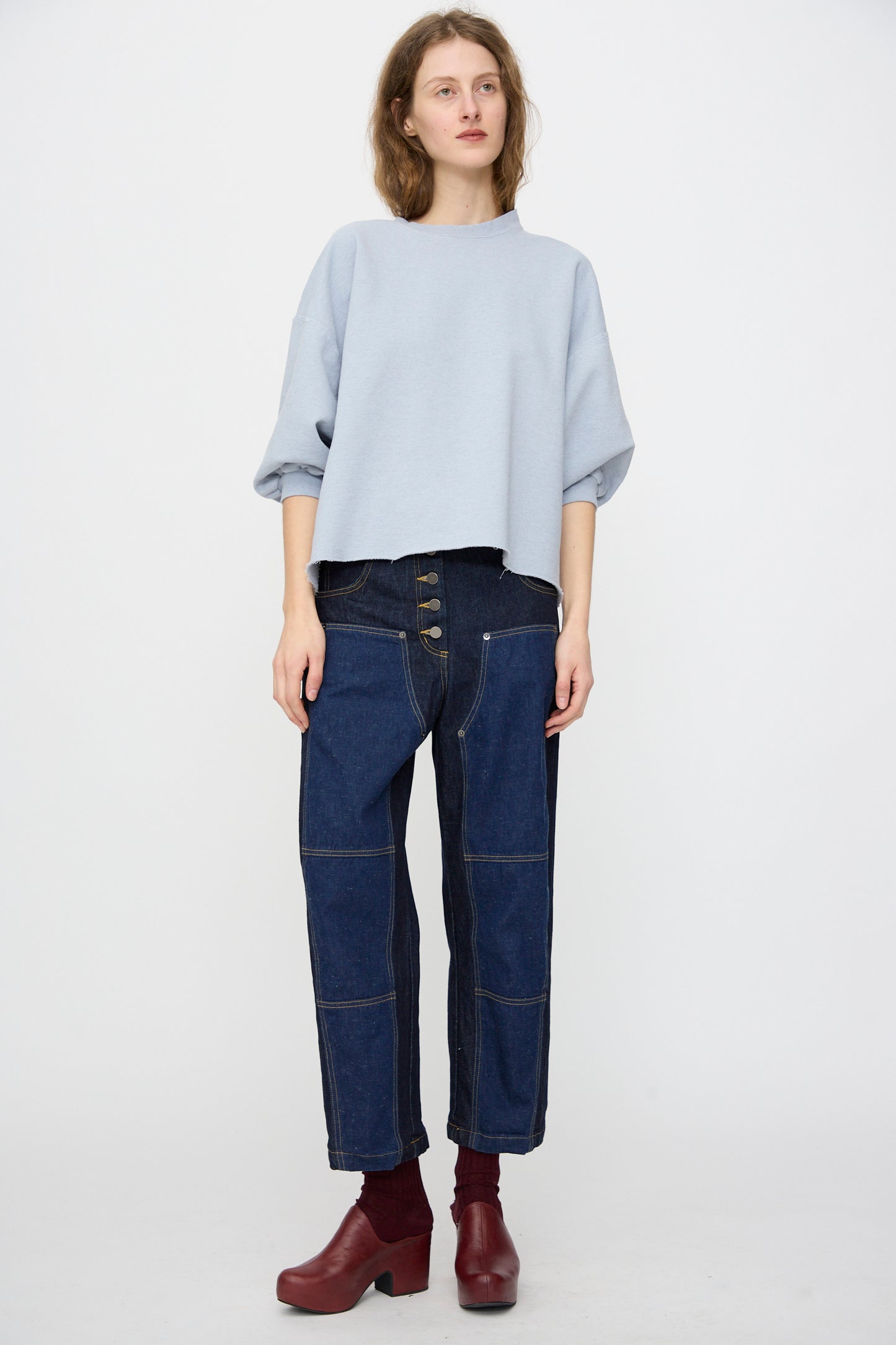 A person wearing the Rachel Comey Fond Sweatshirt in Blue, styled with high-waisted dark blue jeans and maroon heeled boots, stands against a plain white background.