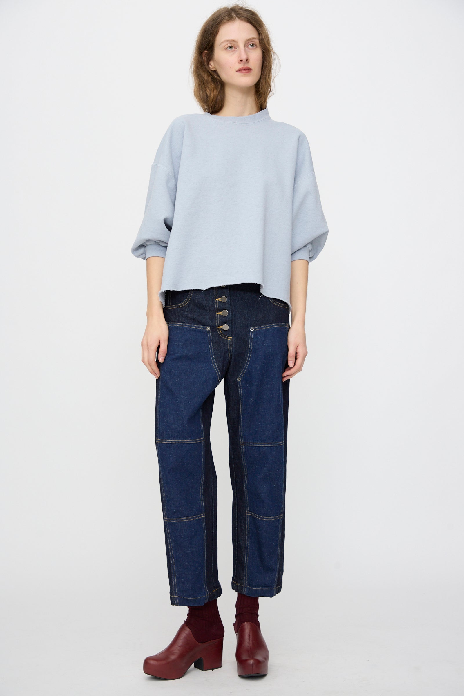 A person wearing the Rachel Comey Fond Sweatshirt in Blue, styled with high-waisted dark blue jeans and maroon heeled boots, stands against a plain white background.