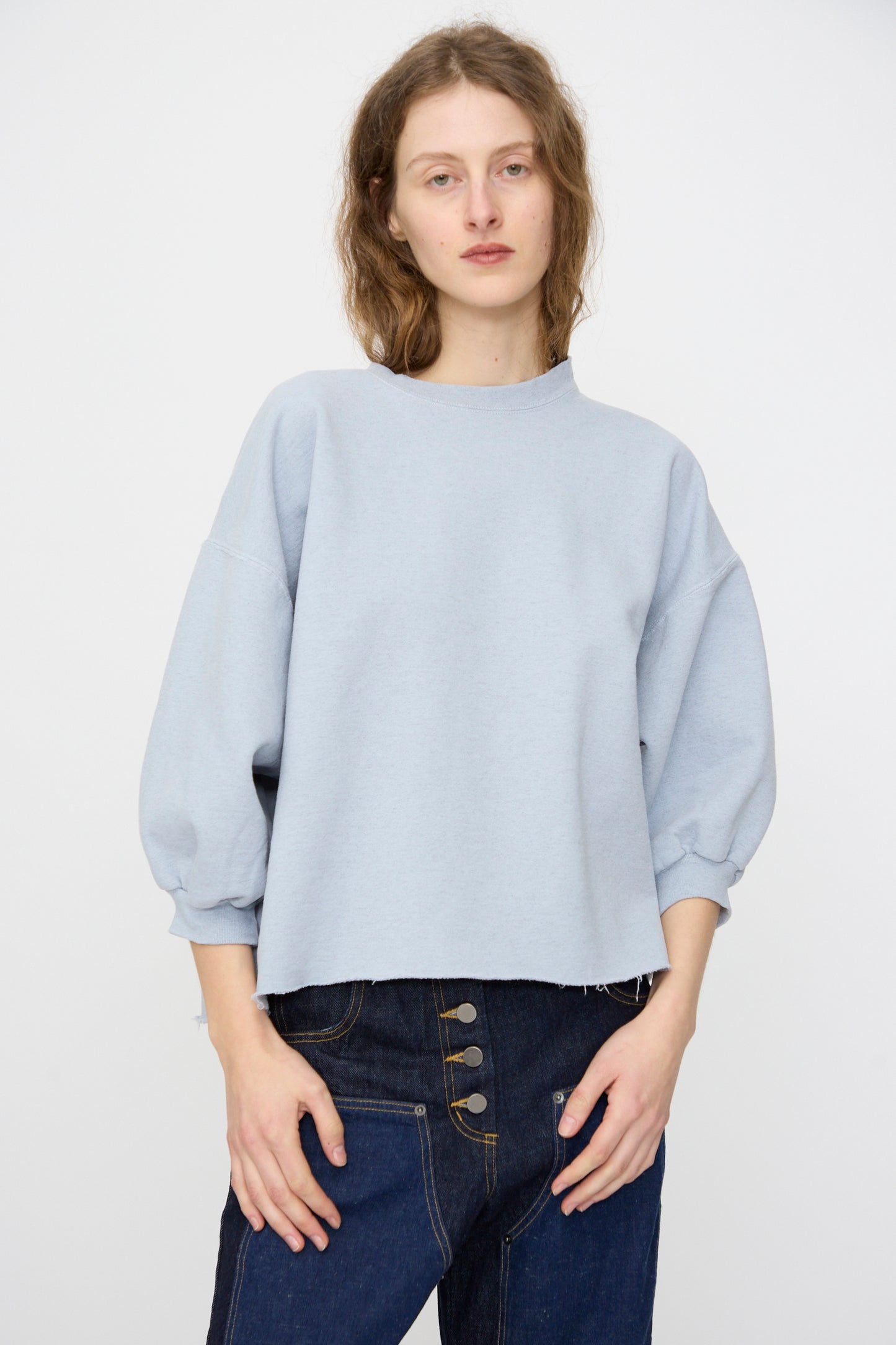 A person wears the Rachel Comey Fond Sweatshirt in Blue—an oversized, genderless style with three-quarter sleeves—paired with high-waisted dark blue jeans featuring a raw edge hemline, standing against a plain background.