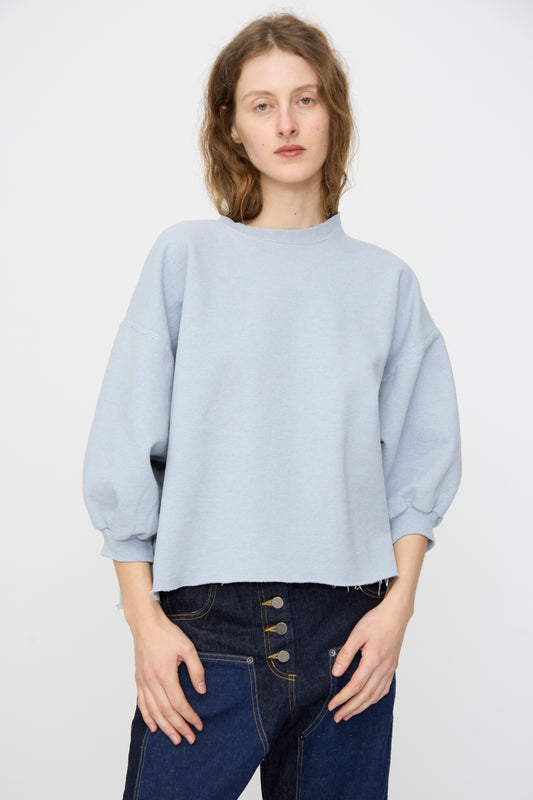 A person wears the Rachel Comey Fond Sweatshirt in Blue—an oversized, genderless style with three-quarter sleeves—paired with high-waisted dark blue jeans featuring a raw edge hemline, standing against a plain background.
