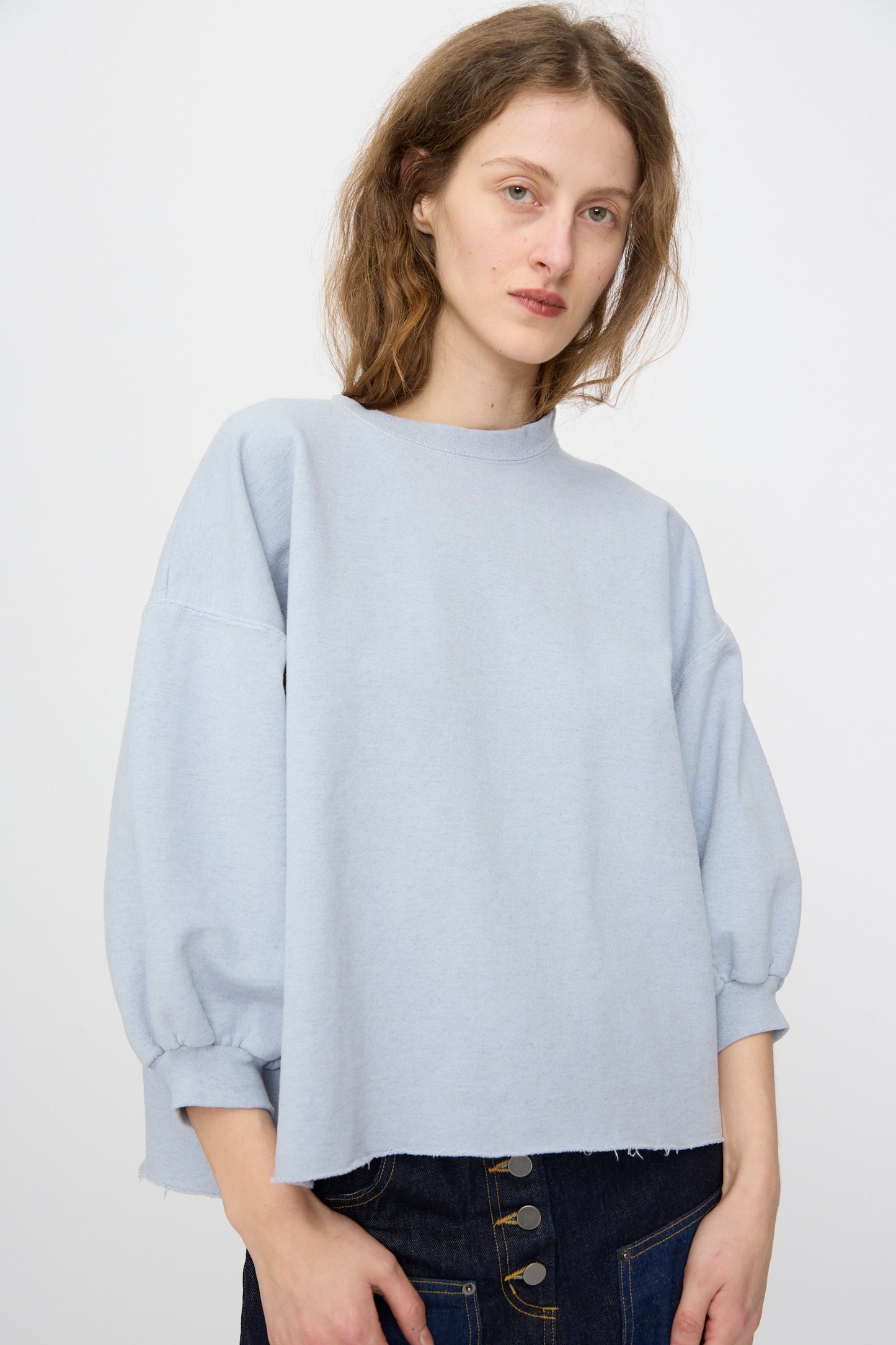 A woman with light brown hair models the Rachel Comey Fond Sweatshirt in blue—a genderless, oversized cotton-blend top—paired with dark high-waisted jeans. She stands against a plain white background.