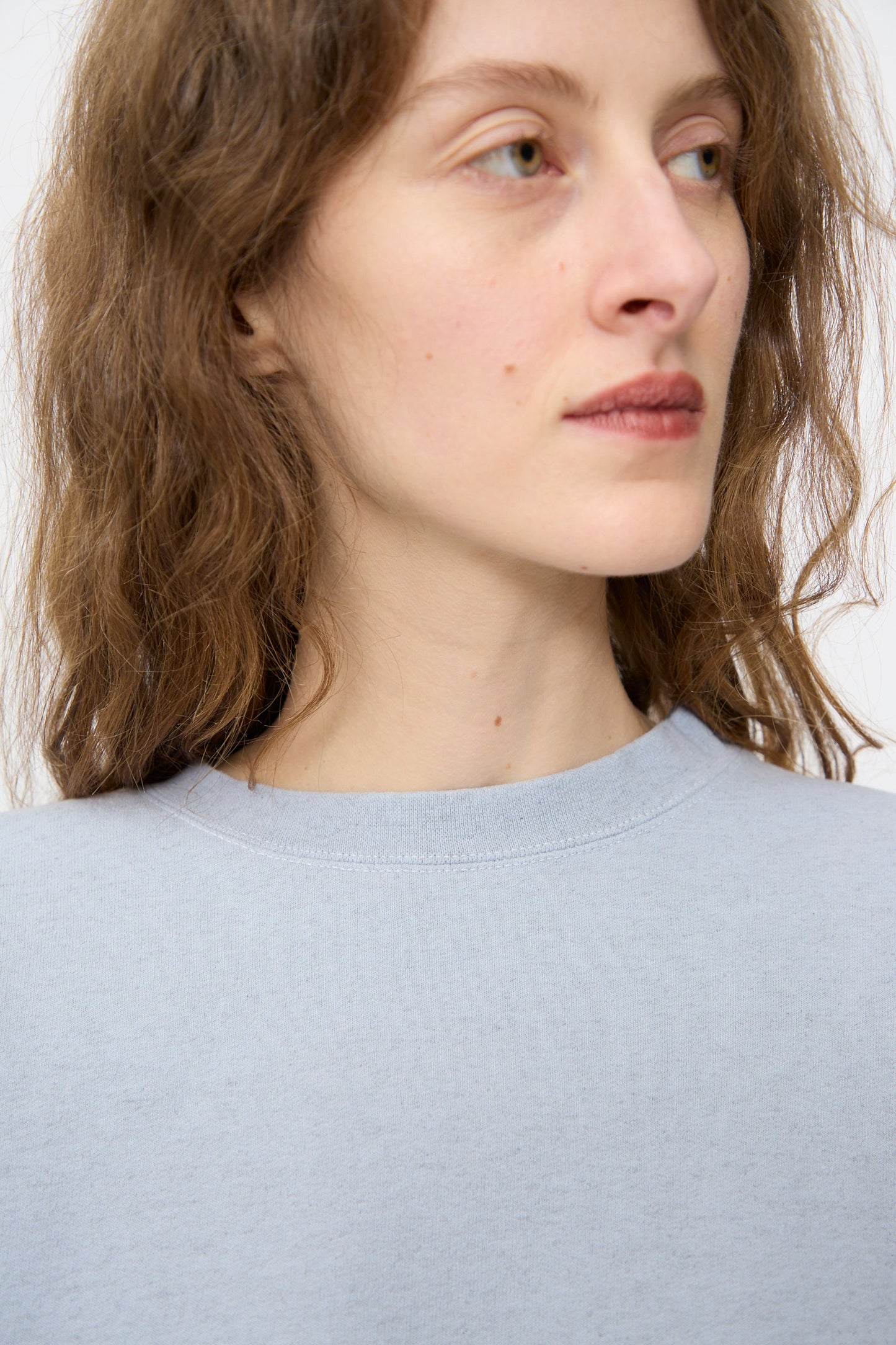 A person with long brown hair wears the Rachel Comey Fond Sweatshirt in Blue—an oversized, genderless piece with a raw edge hemline—while looking slightly to the side against a plain background.