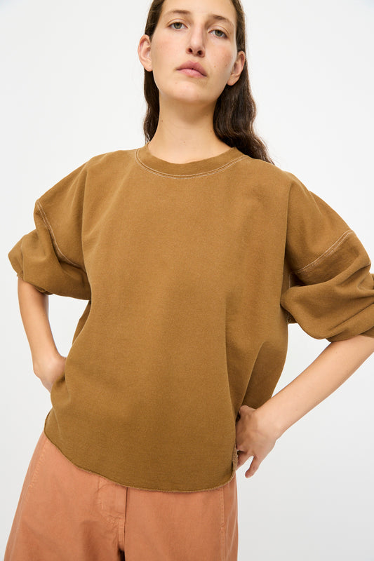 A person wears the Rachel Comey Fond Sweatshirt in Bronze—an oversized, genderless, distressed cotton top with rolled-up sleeves—paired with peach pants, standing against a plain white background.