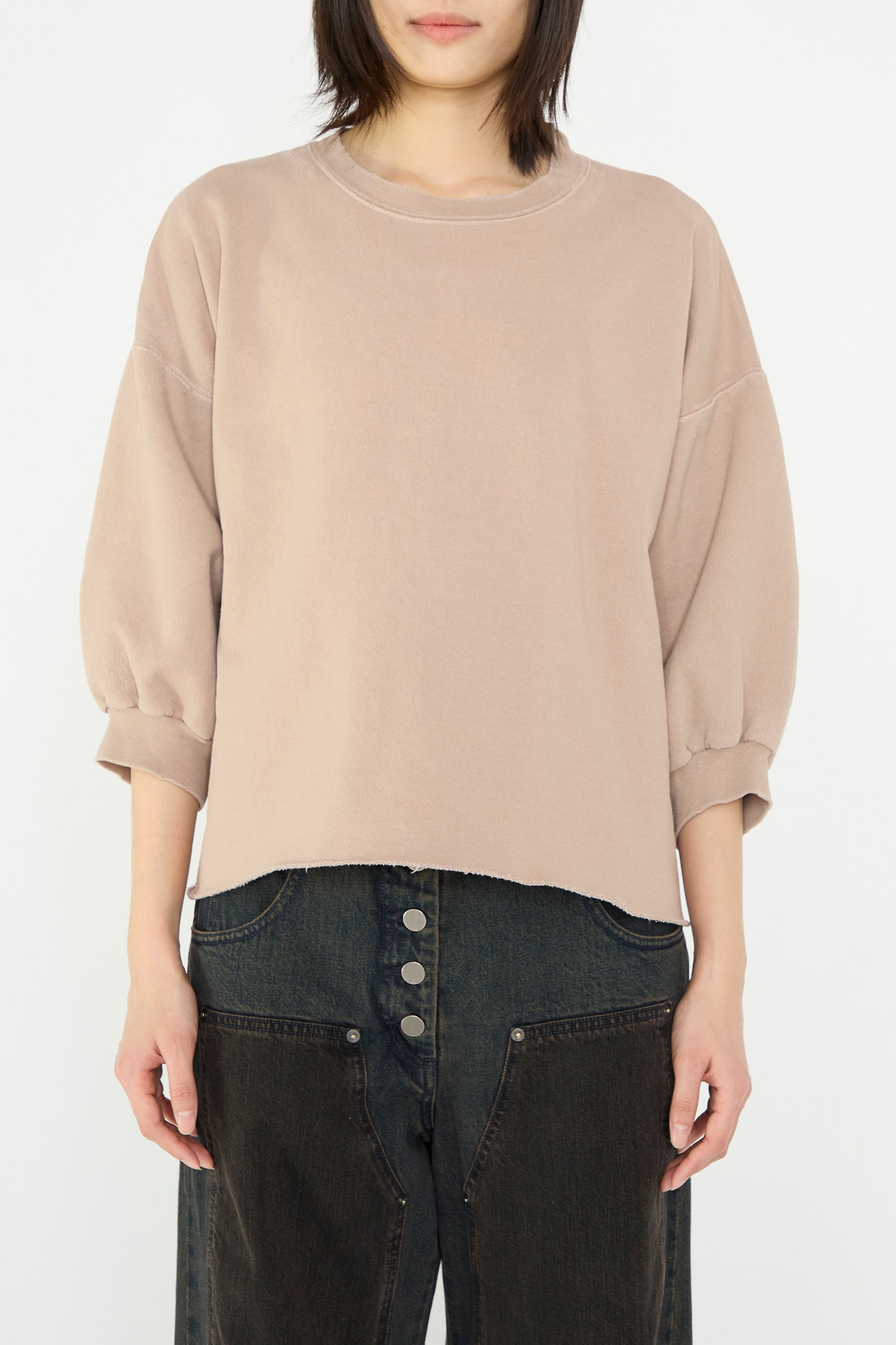 A person in a Rachel Comey Fond Sweatshirt in Buff with cropped sleeves and dark buttoned pants stands against a plain background.