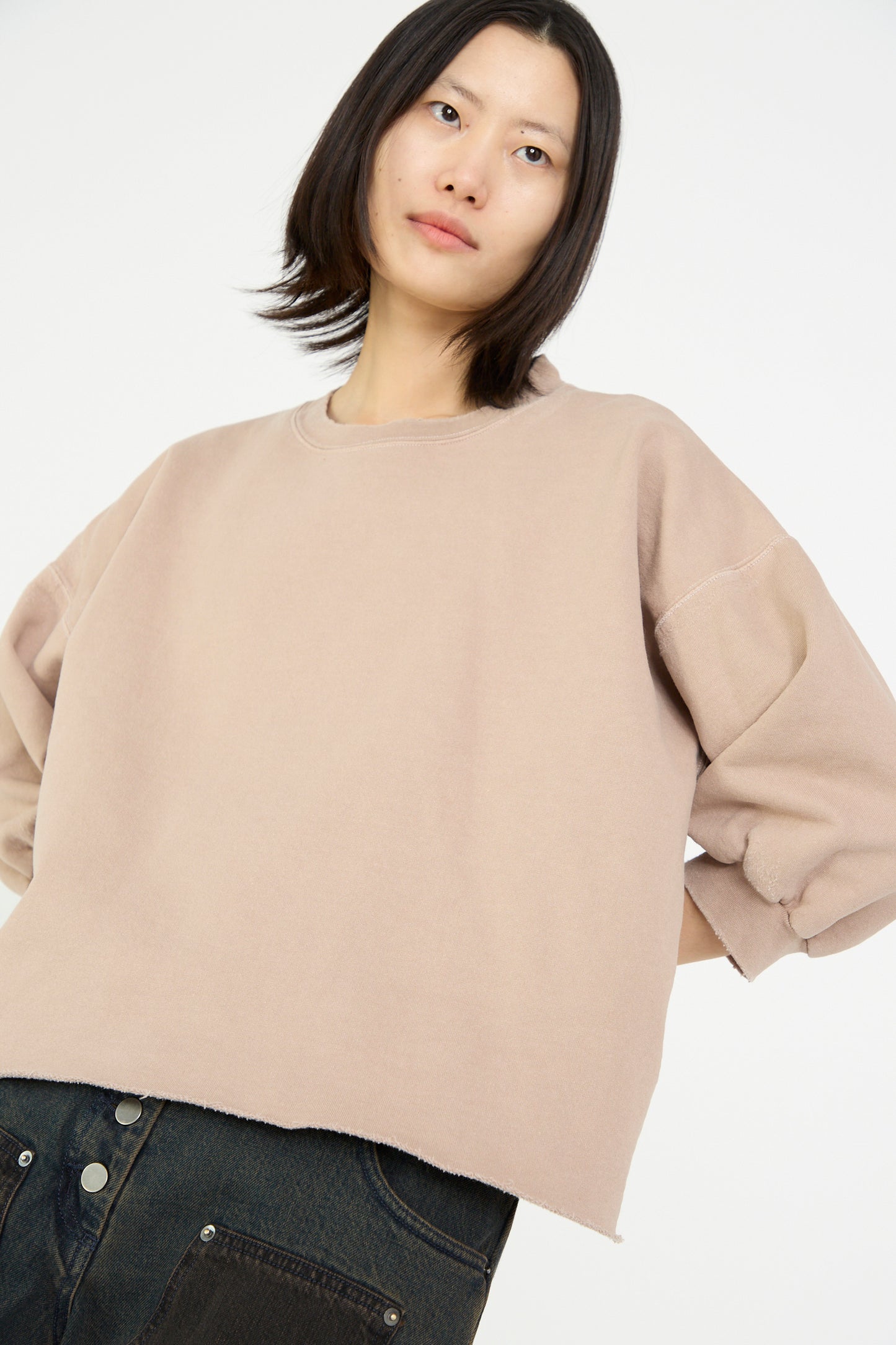 A person with shoulder-length hair wears the Rachel Comey Fond Sweatshirt in Buff, featuring an oversized crewneck and cropped sleeves, paired with dark jeans while glancing to the side against a plain background.