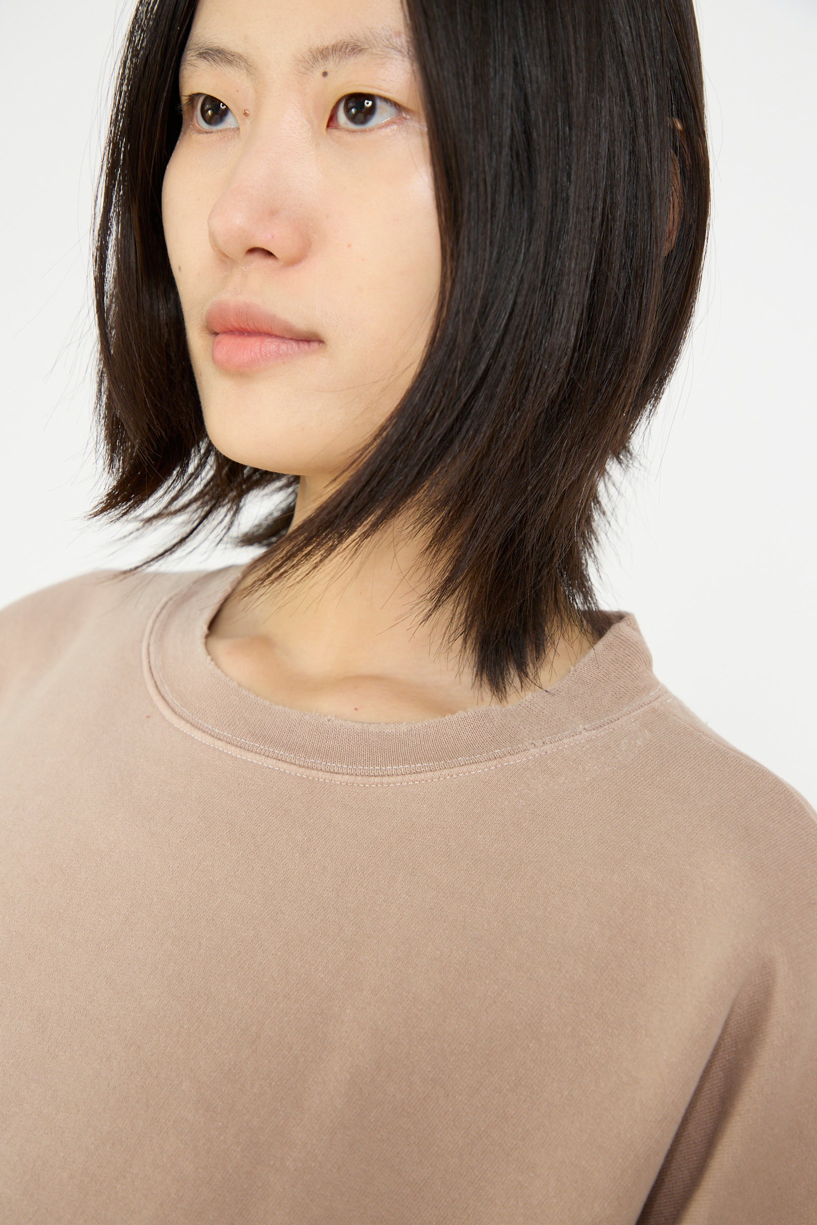 A person with shoulder-length hair wears a Fond Sweatshirt in Buff by Rachel Comey, featuring cropped sleeves, and glances to the side against a plain background.