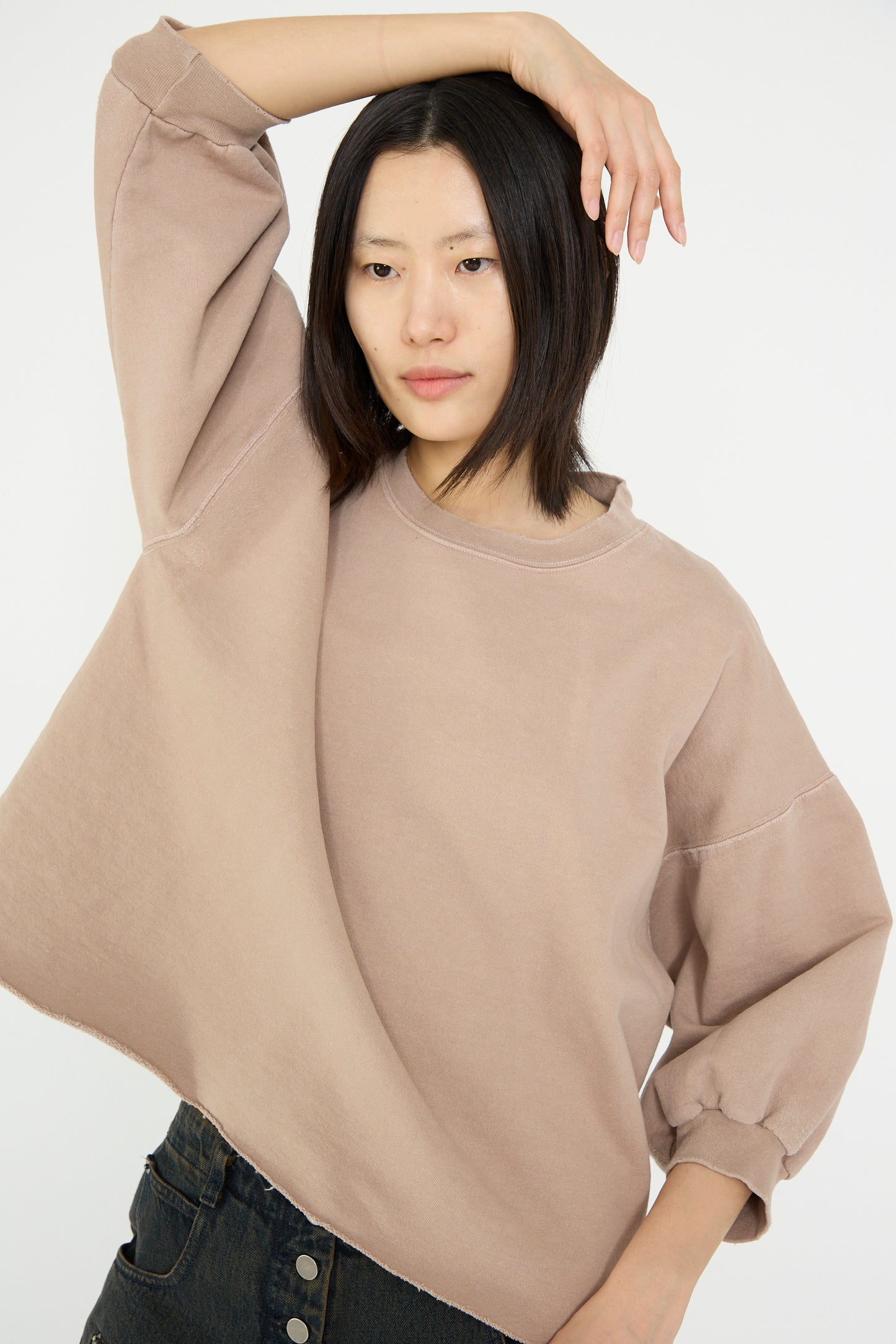 A model wearing the Rachel Comey Fond Sweatshirt in Buff and dark jeans poses with one arm raised against a plain background. 