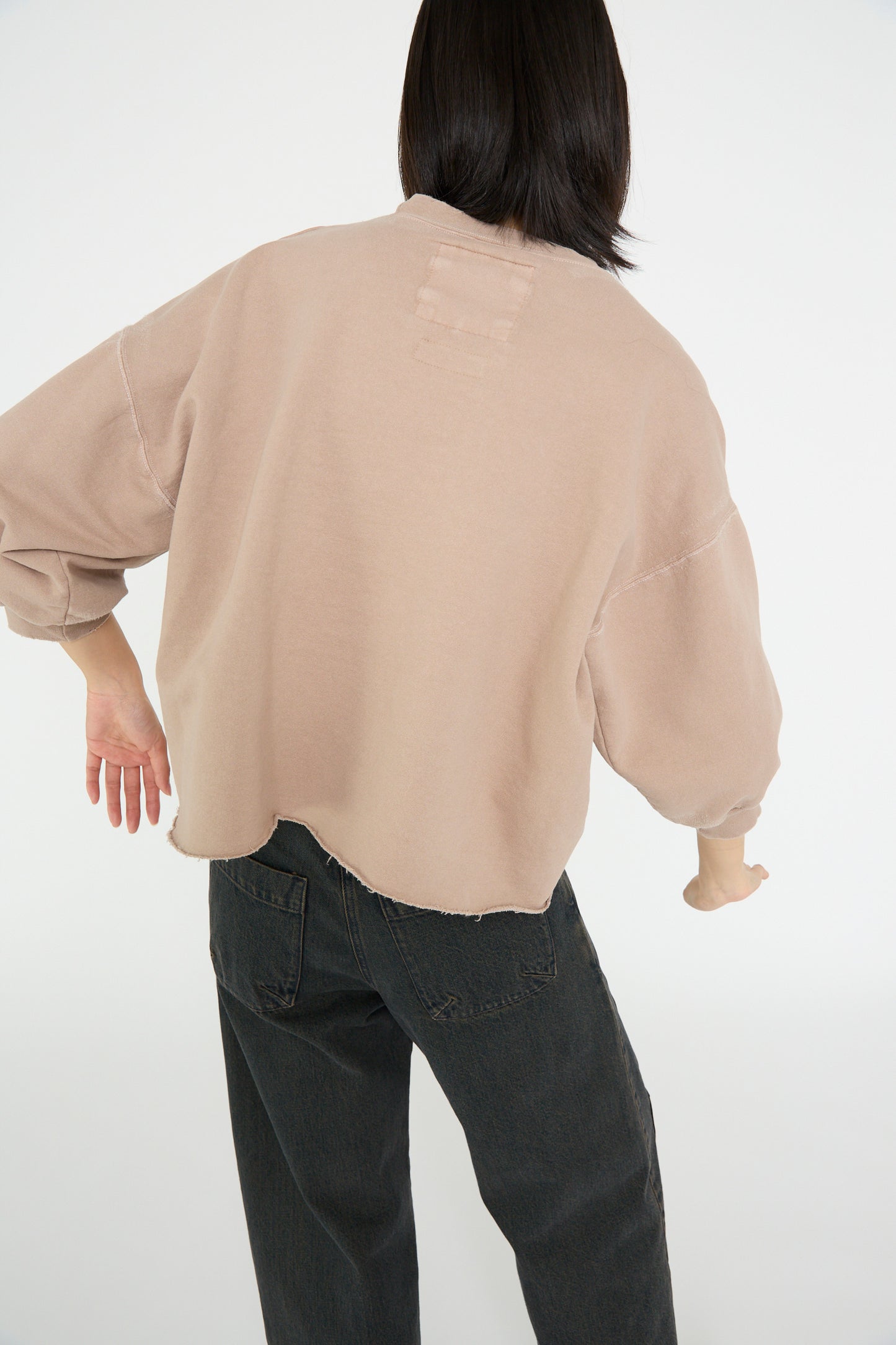 With shoulder-length dark hair, a person is wearing the Rachel Comey Fond Sweatshirt in Buff with cropped sleeves and black jeans, facing away with their left arm slightly raised.